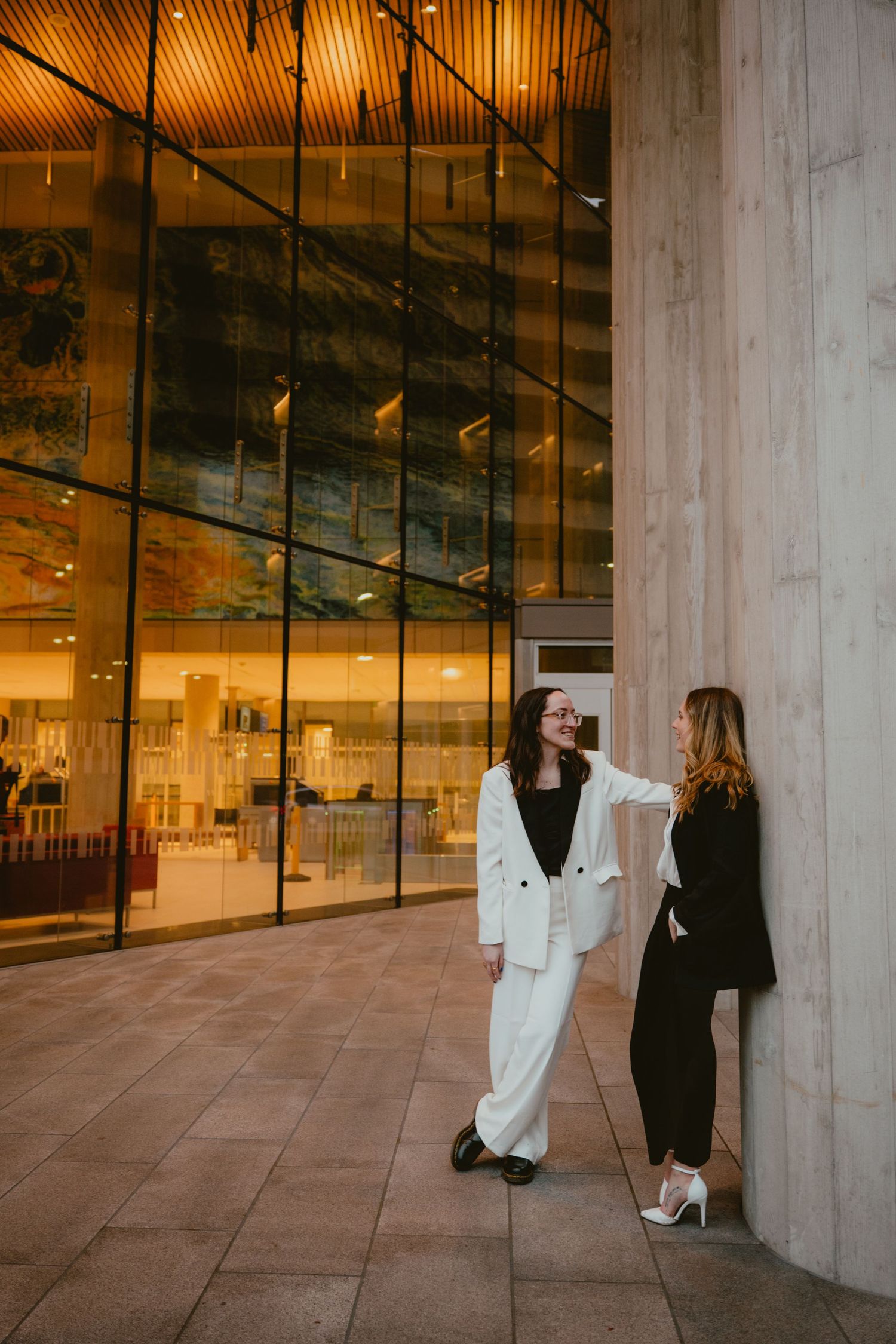 Two women in black and white suits standing and smiling outside a glass building.