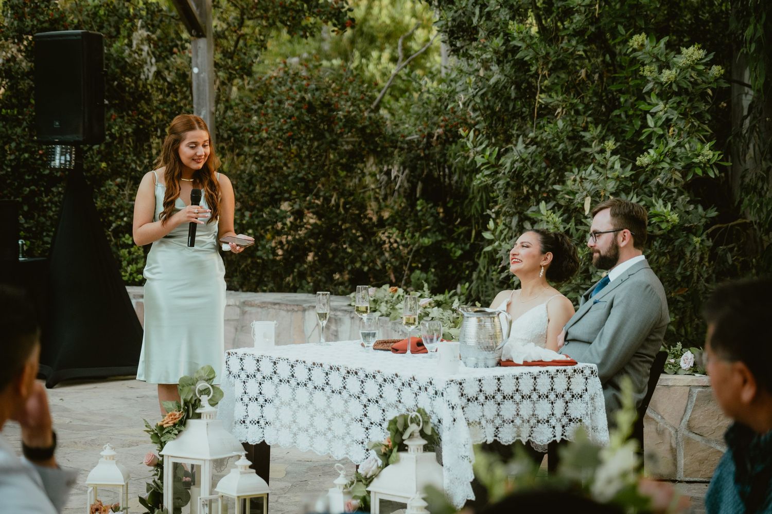 Bridesmaid delivers heartfelt speech to smiling bride and groom at outdoor reception.