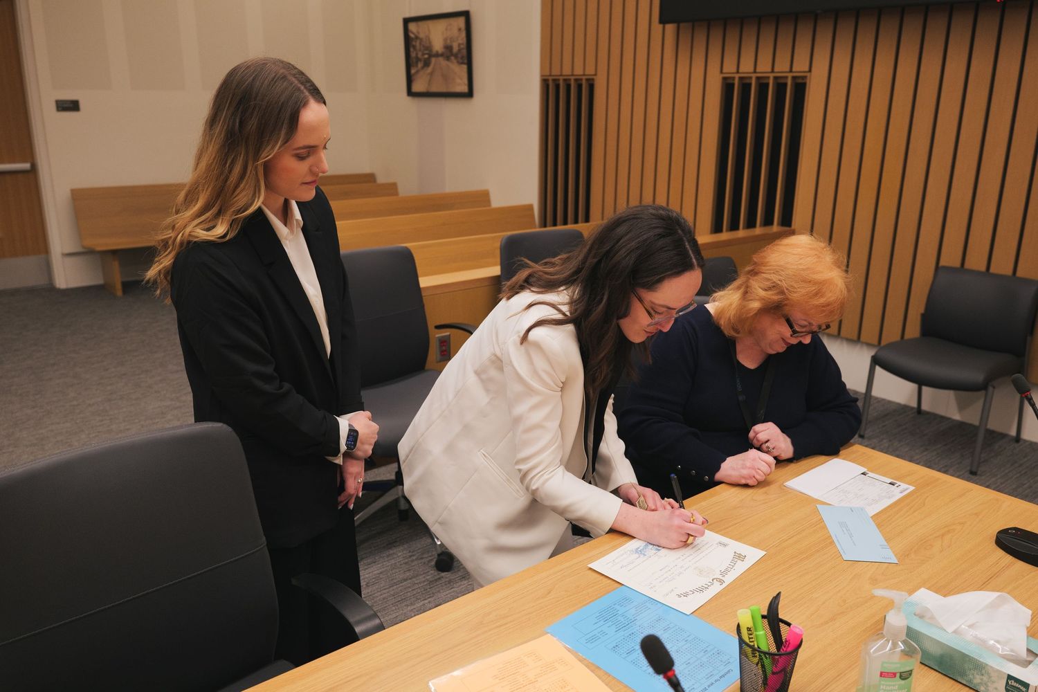 Three women review documents at a courtroom table in a wood-paneled legal setting.