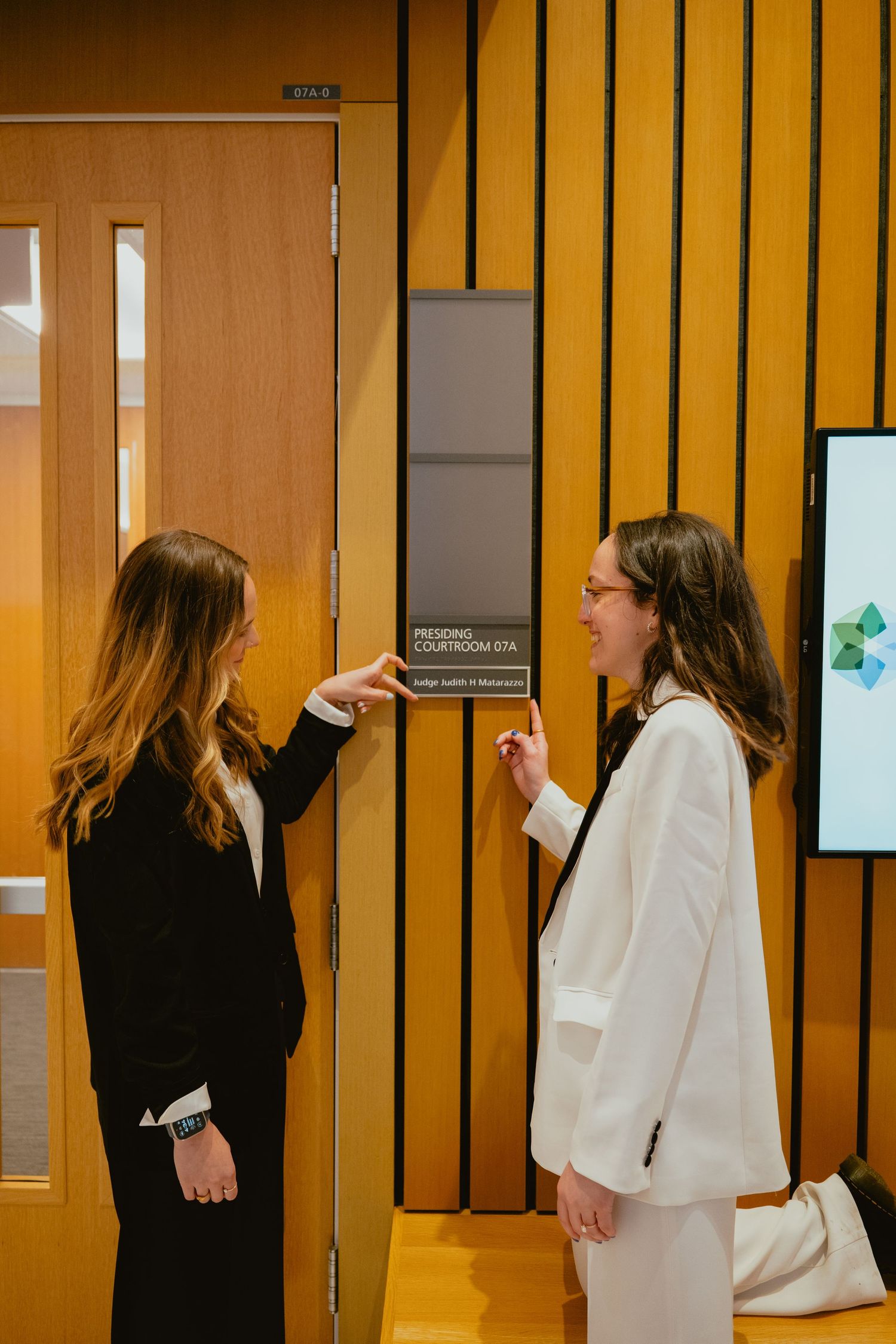 Two women in professional attire stand outside a presiding courtroom door in a law building.