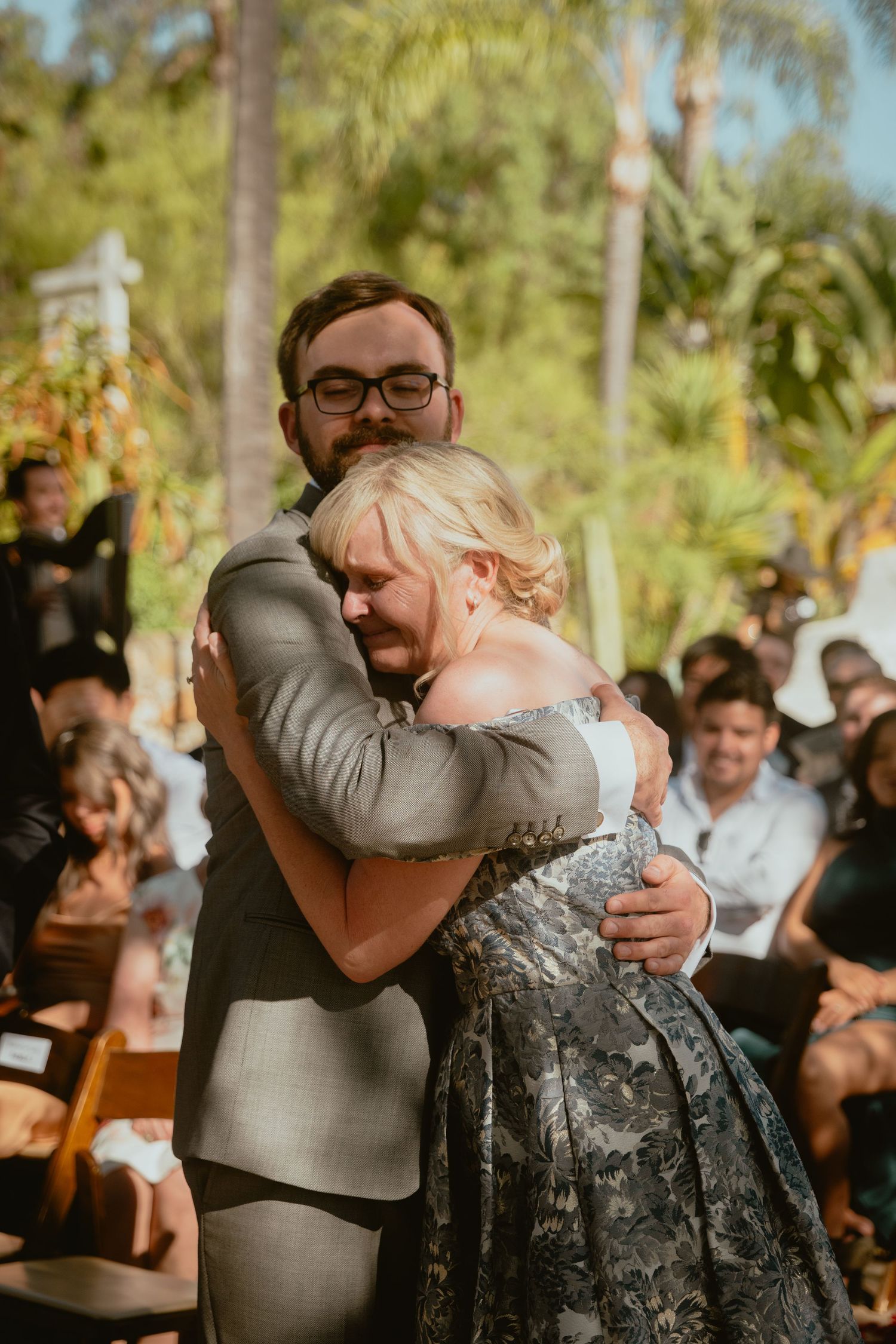 Groom in grey suit shares a heartfelt embrace with an emotional blonde woman at the ceremony.