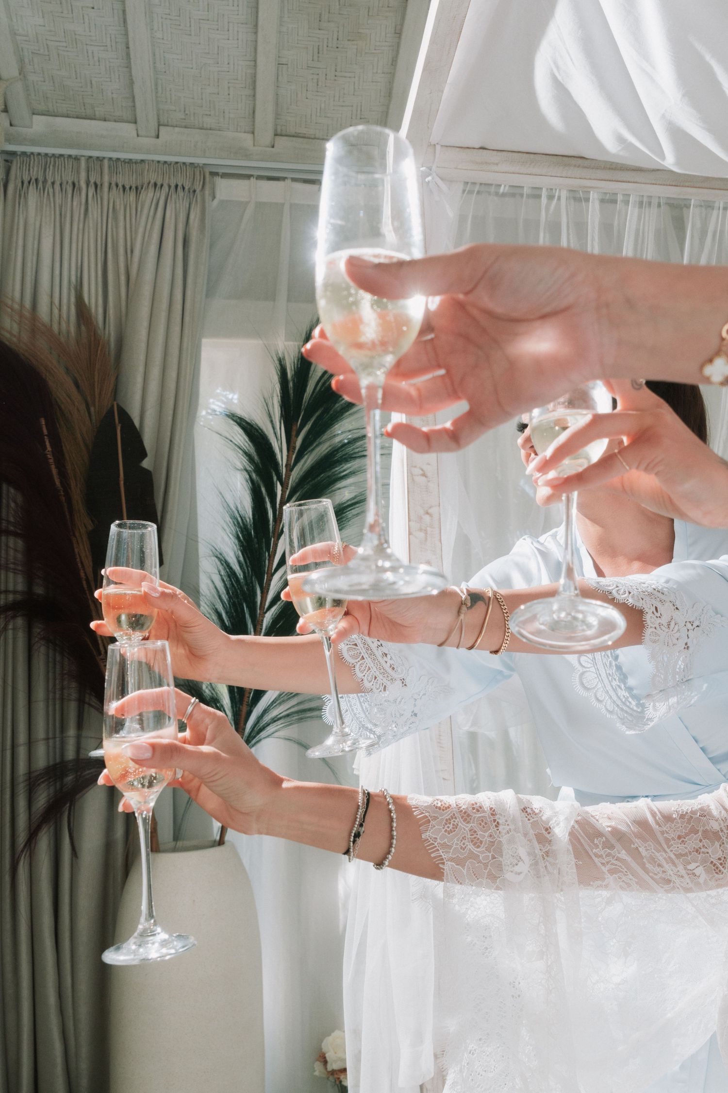 Bridal party clinks champagne glasses in sunlit room during wedding morning preparations.