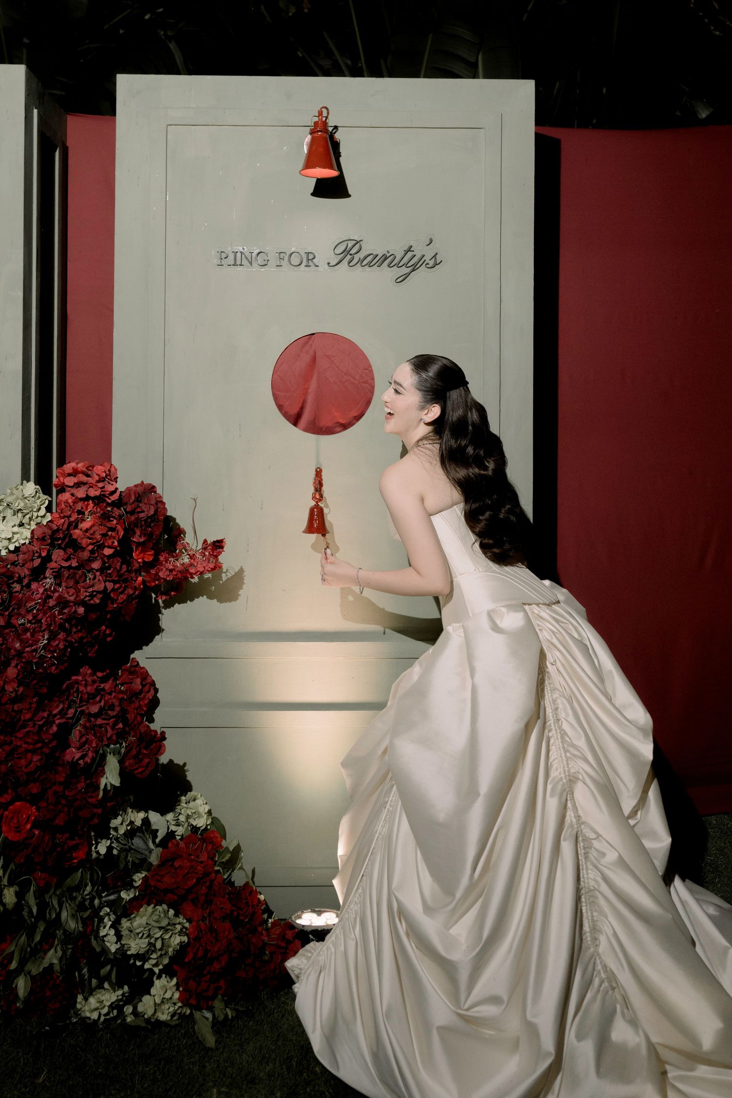 Bride in strapless white ball gown ringing a red bell at an elegant wedding reception with red floral decor.