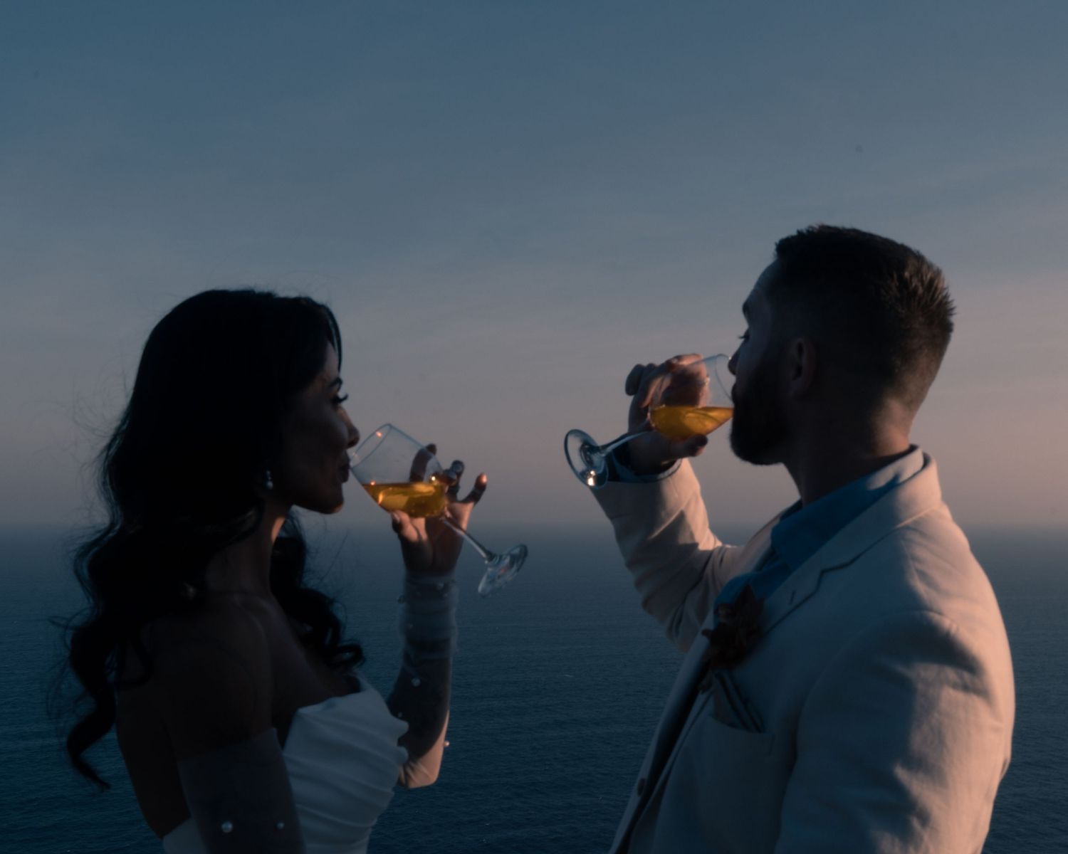 Couple silhouetted against ocean twilight sky toasting with glasses of white wine at sunset.