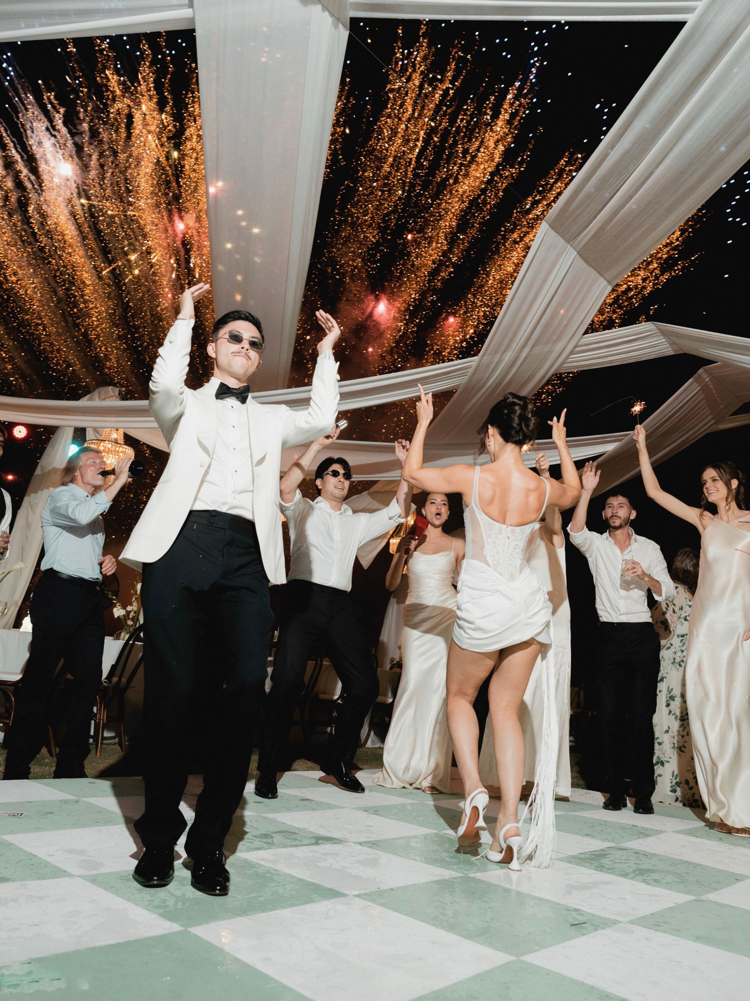 Bride and groom celebrate on dance floor with fireworks and cheering wedding guests.