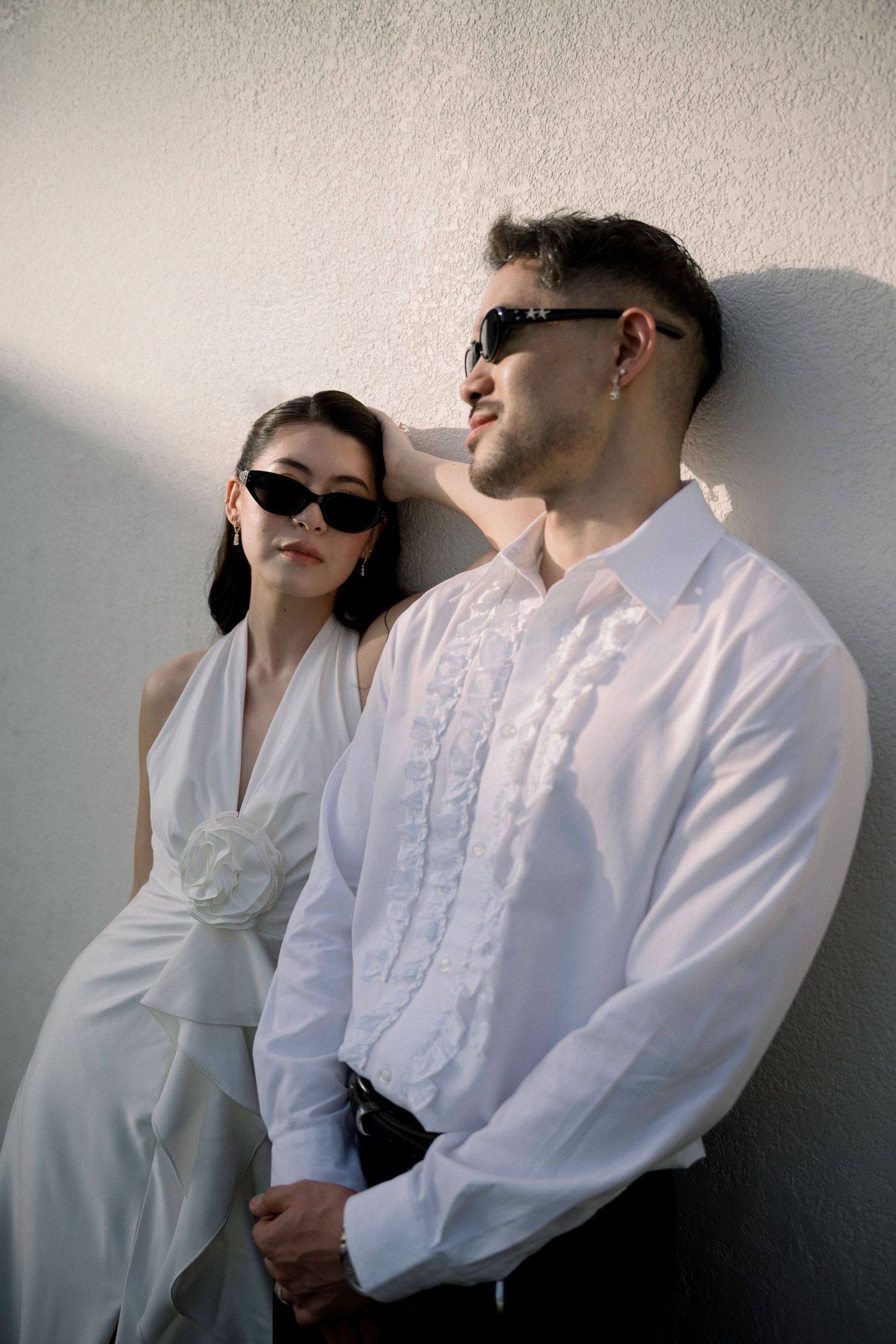 Stylish couple in white outfits and sunglasses lean against a sunlit white wall.