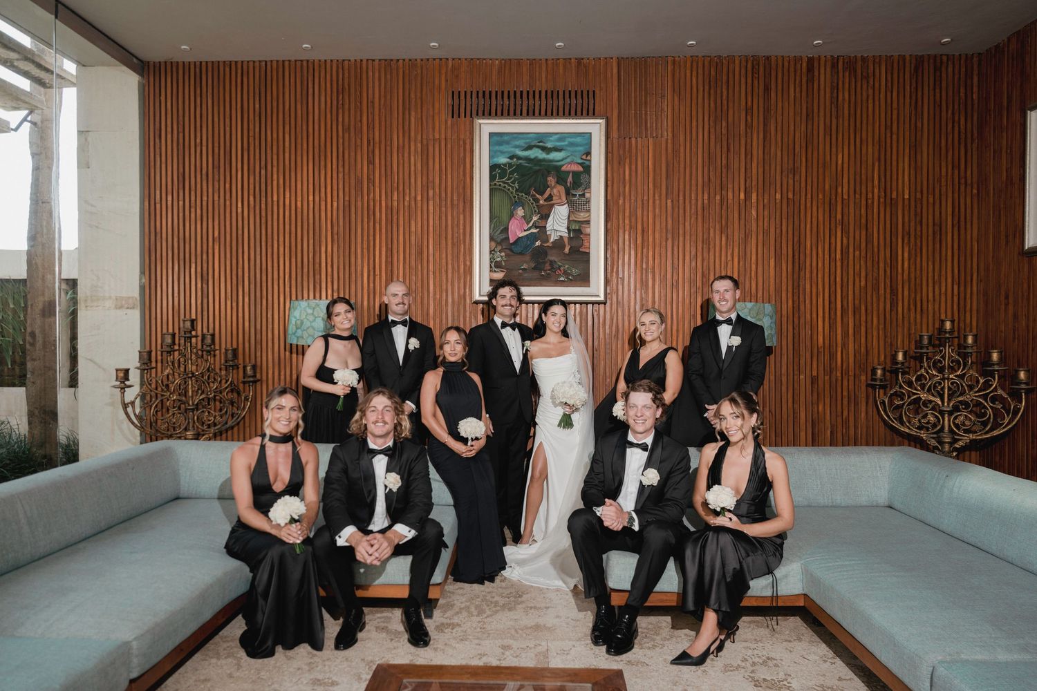 Wedding party group portrait in modern wood-paneled interior with bride in white and guests in black formal wear.