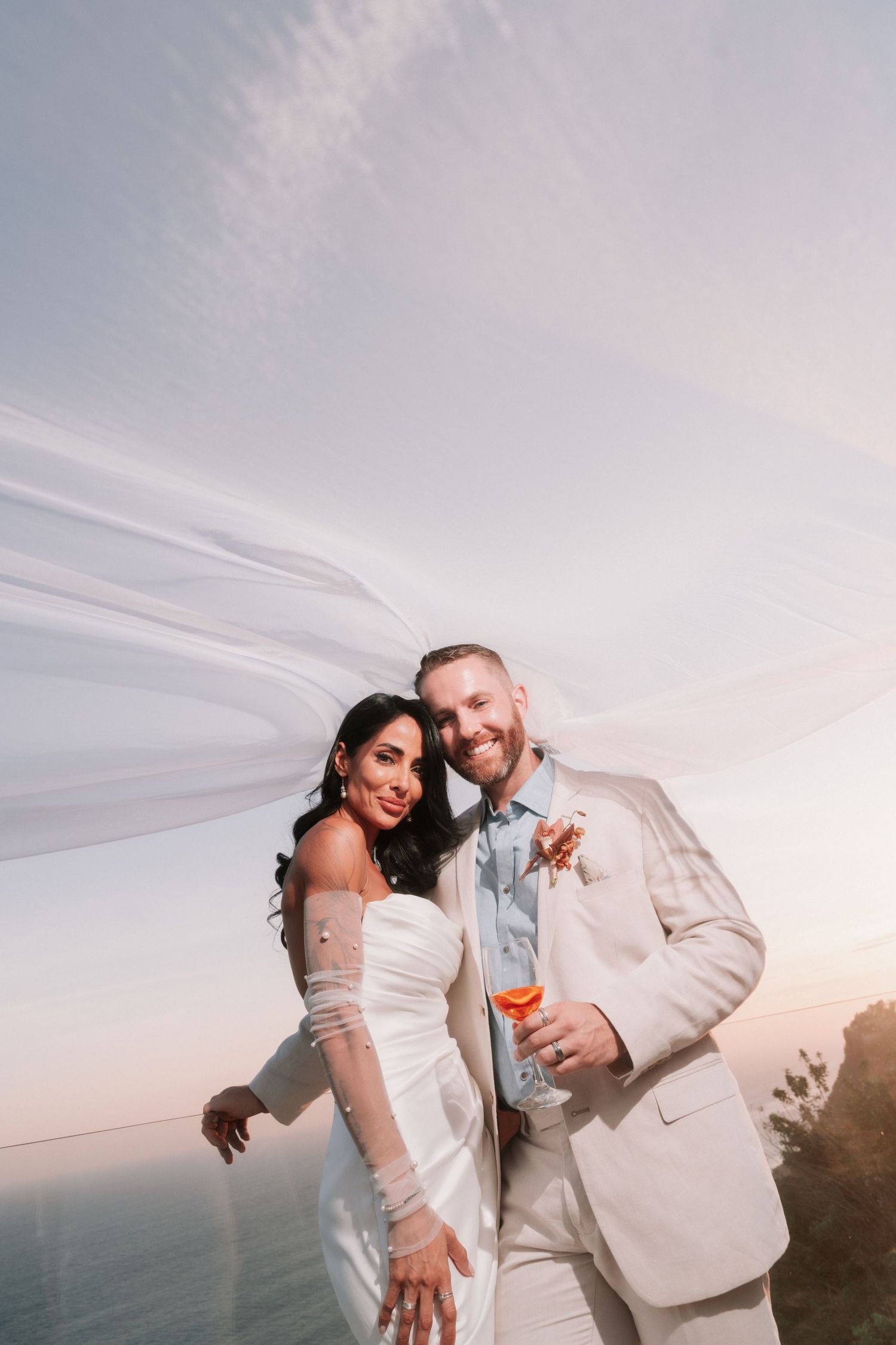 Smiling bride and groom in white attire pose with flowing veil against a dramatic sunset sky.