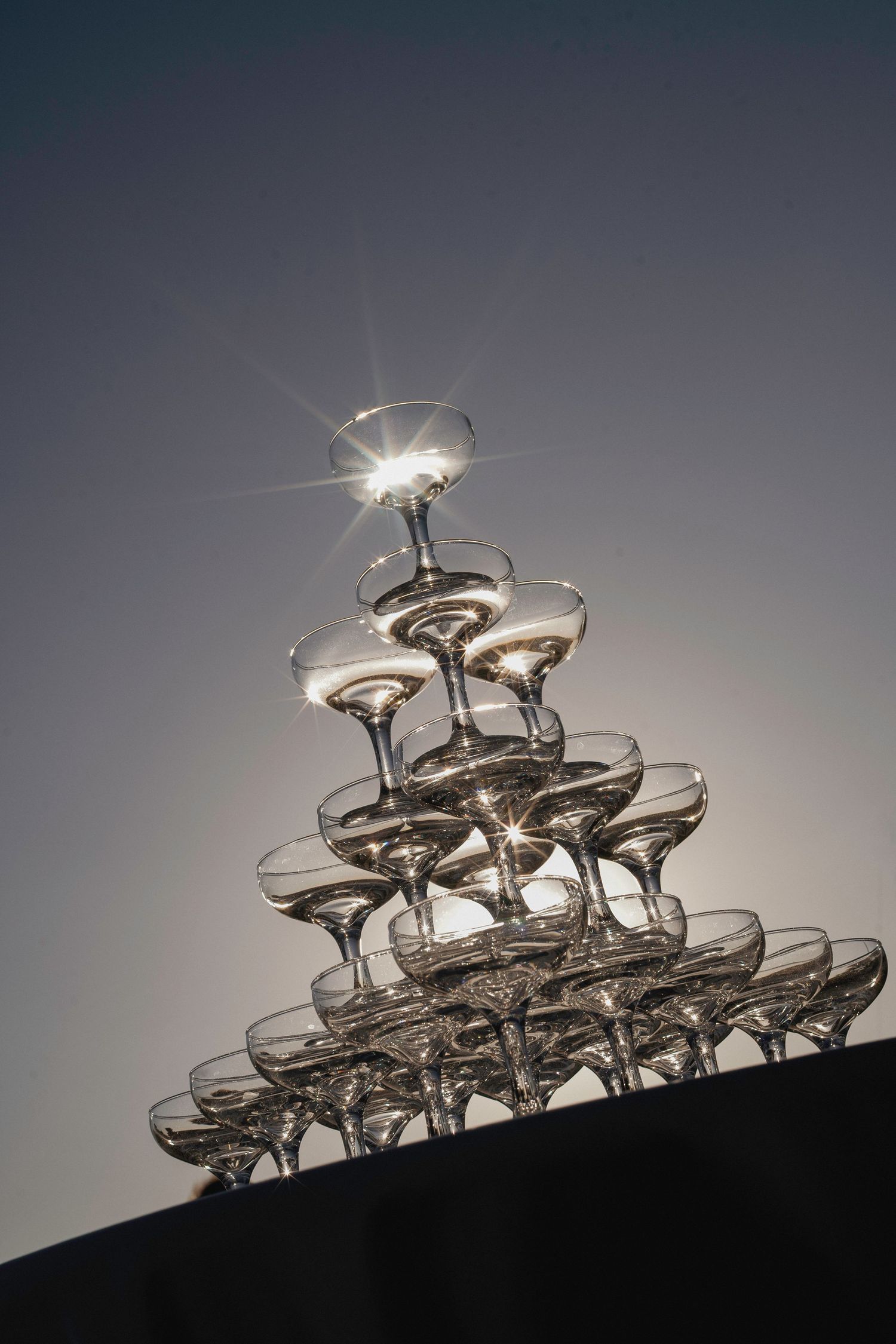 Elegant pyramid tower of champagne coupe glasses backlit by sunlight at wedding reception.