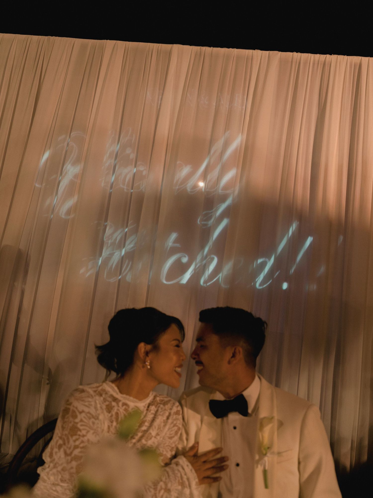 Couple shares romantic moment under glowing projected names on draped backdrop at wedding reception.