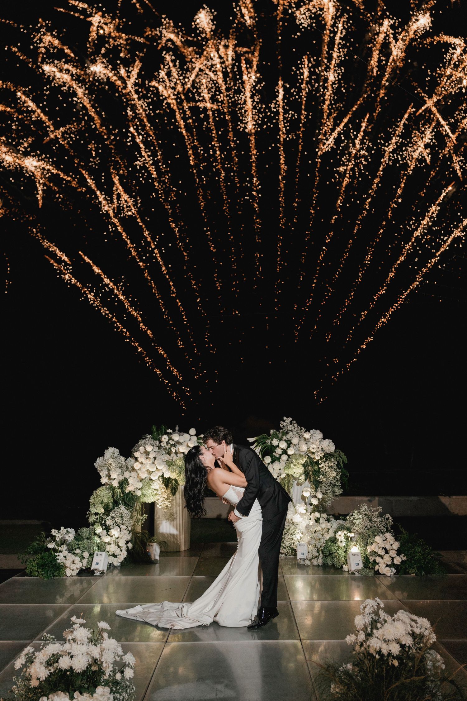 Bride and groom kissing under spectacular fireworks display surrounded by white floral arrangements at night.