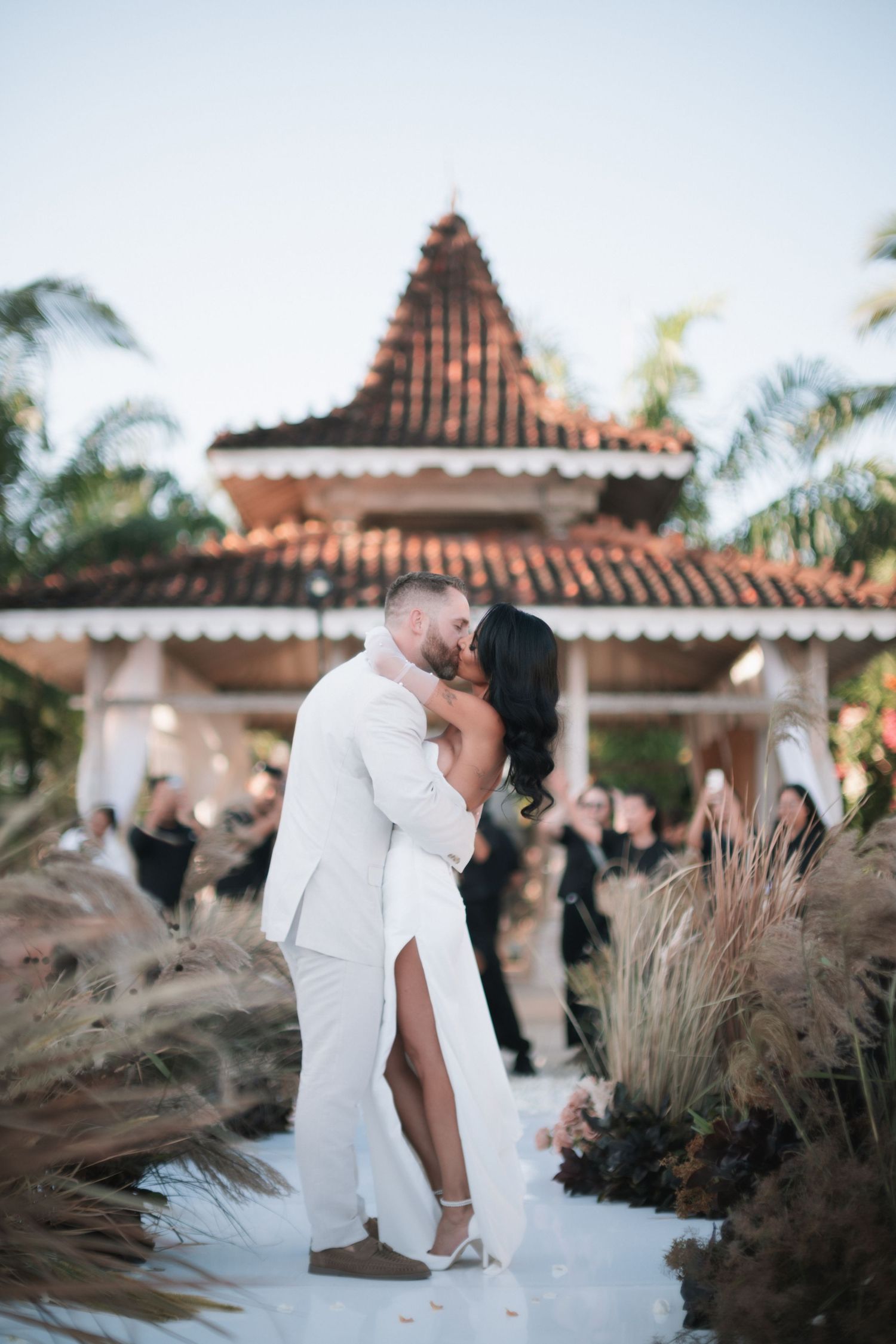 Bride and groom share first kiss on white aisle surrounded by pampas grass at outdoor ceremony.