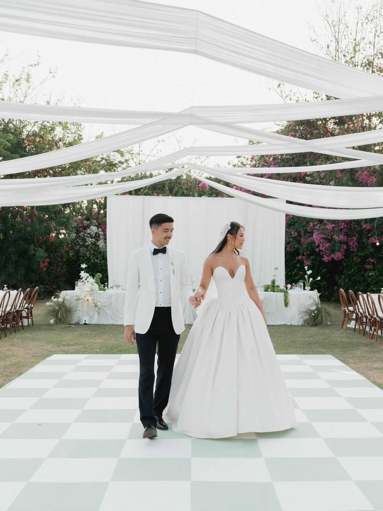 Bride and groom walk on checkered dance floor under white draped canopy at outdoor garden wedding.