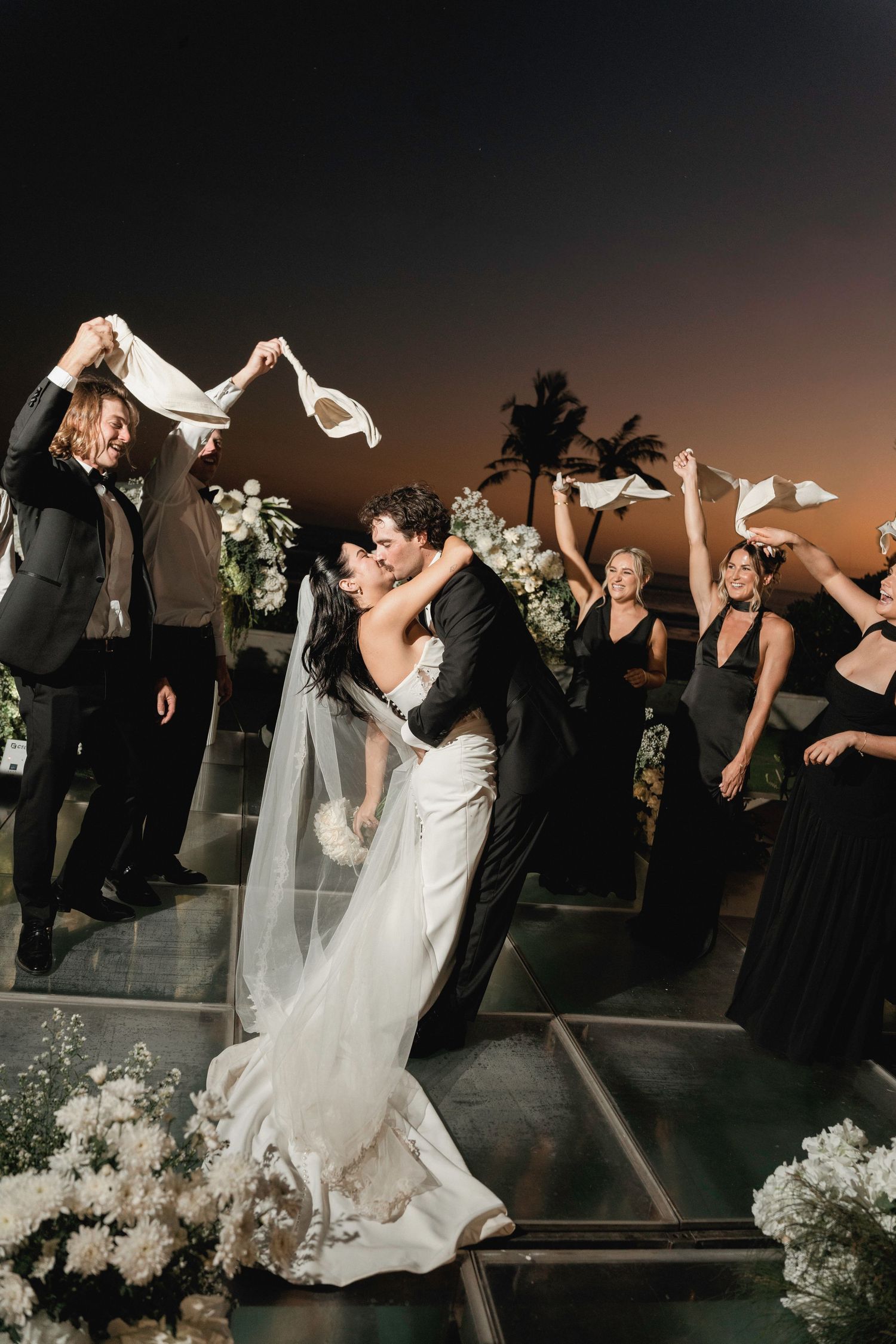 Bride and groom sharing dramatic dip kiss at altar while wedding party waves white handkerchiefs at night.
