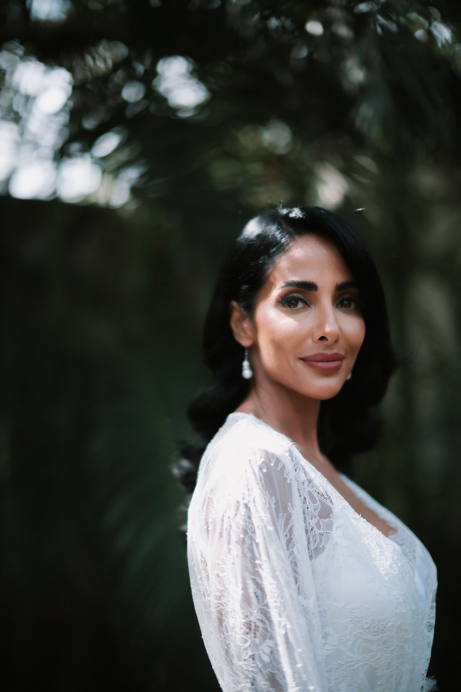 Beautiful dark-haired bride in white lace dress poses outdoors among green foliage.