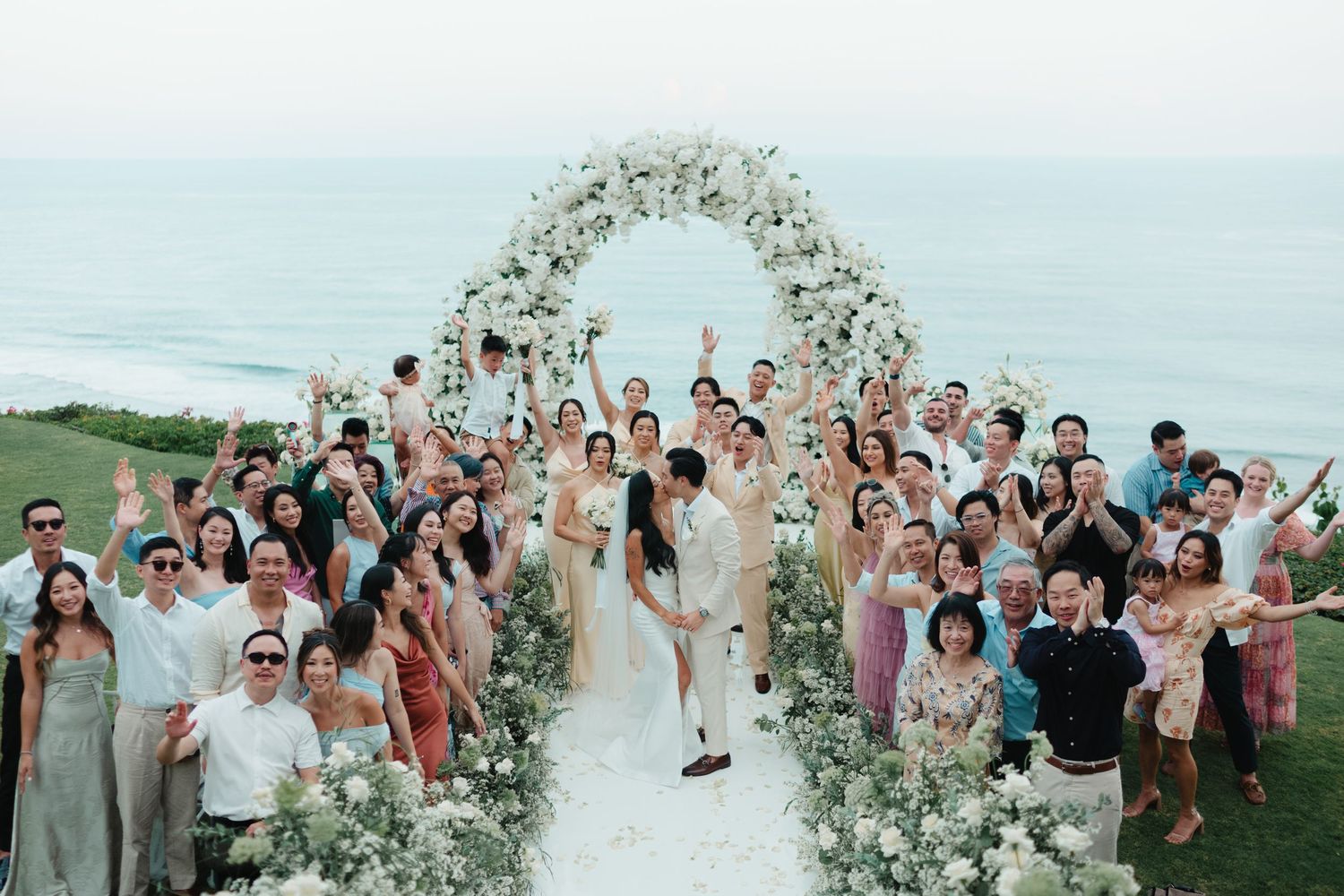 Joyful wedding party celebrating with couple under floral arch with ocean backdrop in Bali.