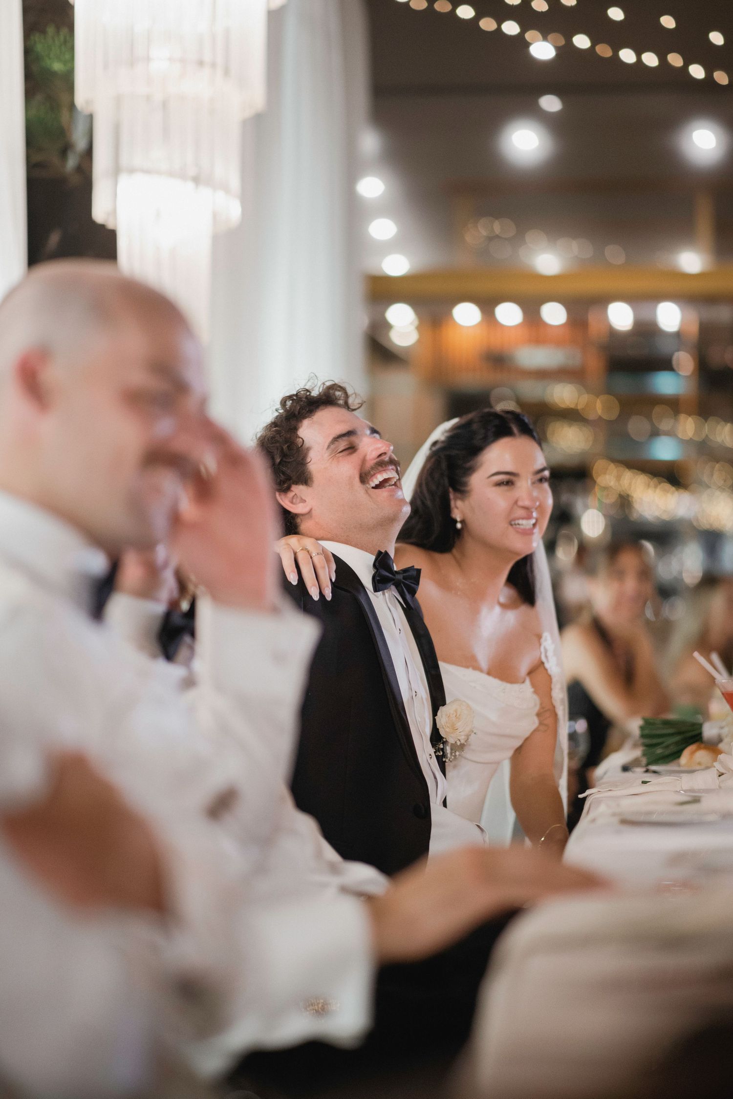 Bride and groom laughing joyfully at wedding reception dinner table with bokeh lights in background.
