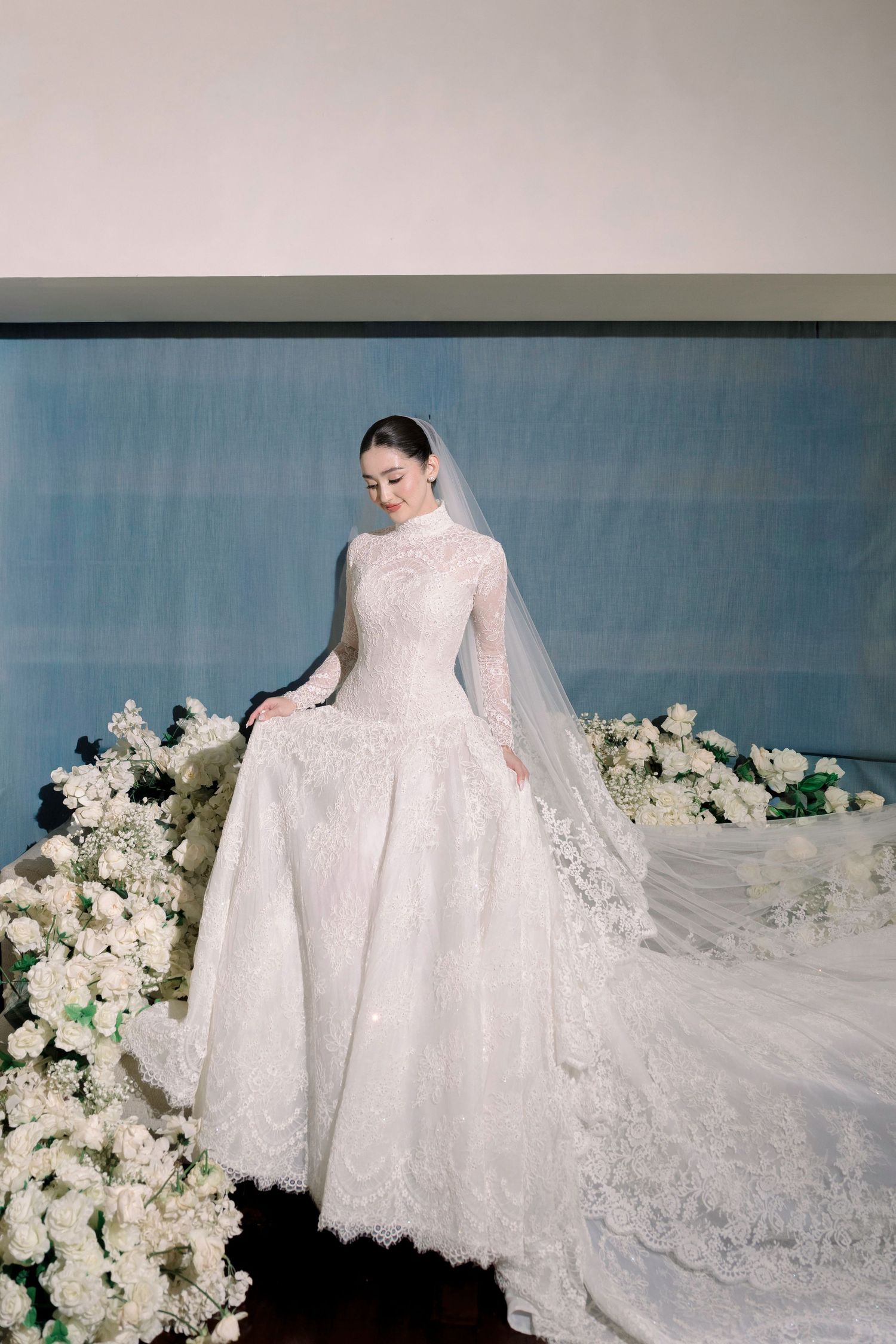 Bride in elegant high-neck long-sleeve lace wedding gown with cathedral train surrounded by white flowers.
