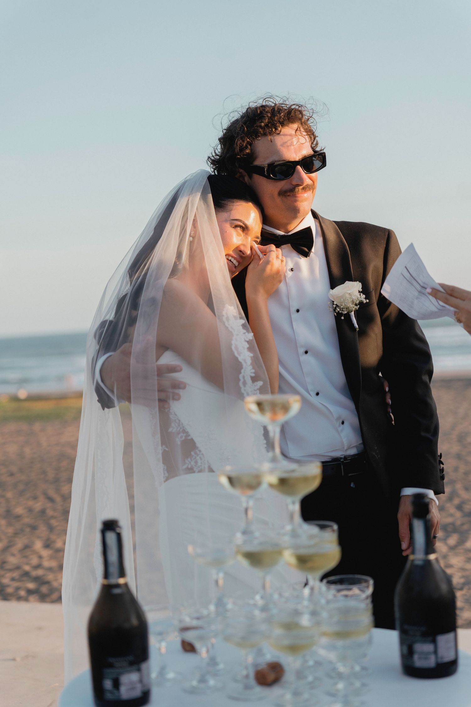 Bride and groom laughing by champagne tower on beach at sunset, groom wearing sunglasses and black tuxedo.