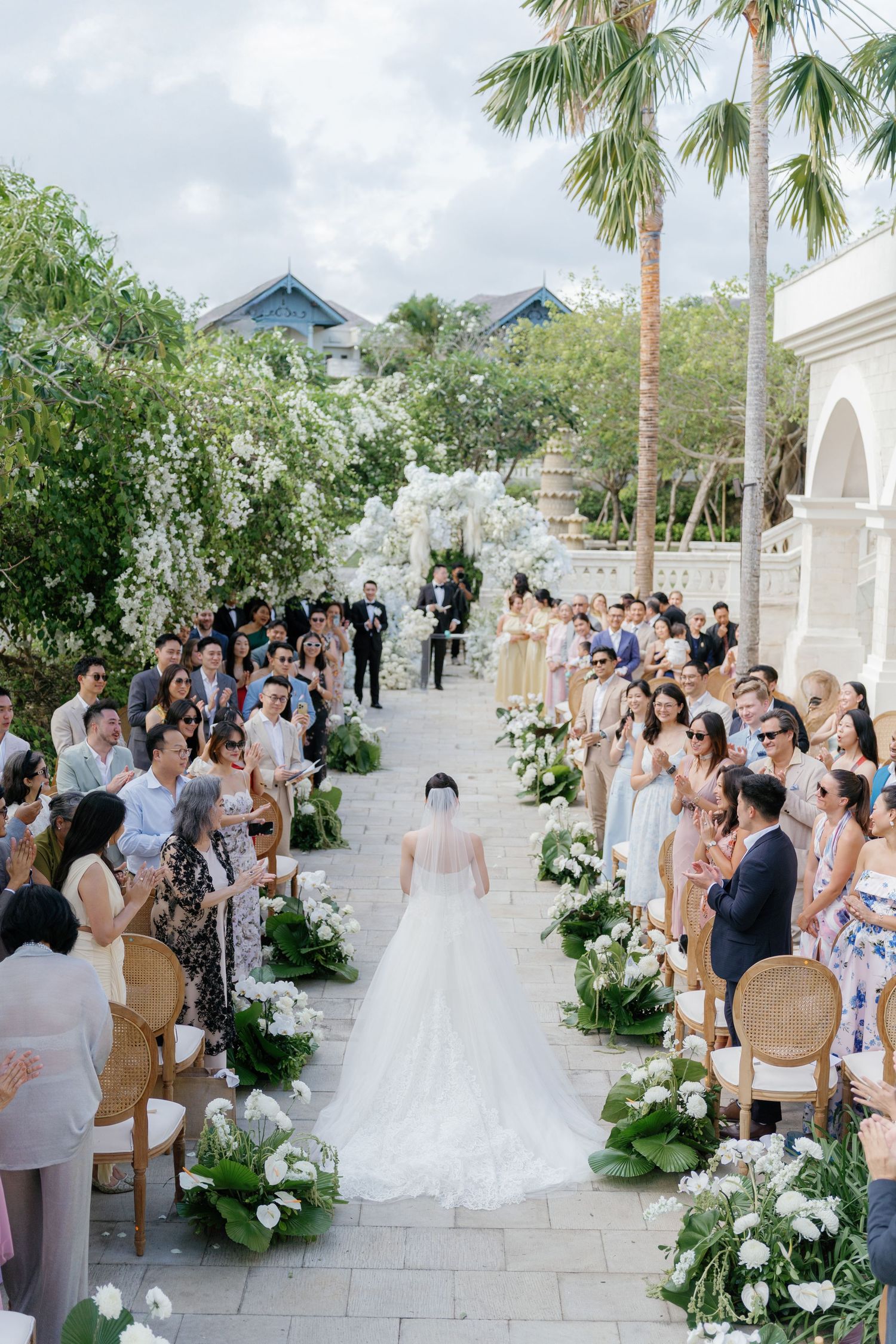 Bride walks down floral aisle at elegant outdoor wedding ceremony with guests seated among white blooms and palm trees.