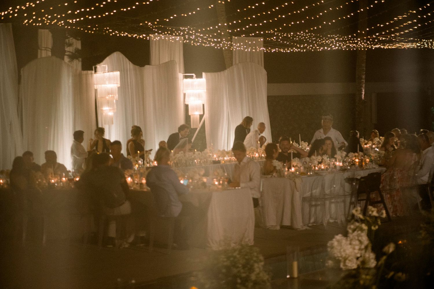 Romantic candlelit wedding reception under string lights with white draping and guests dining at long tables.