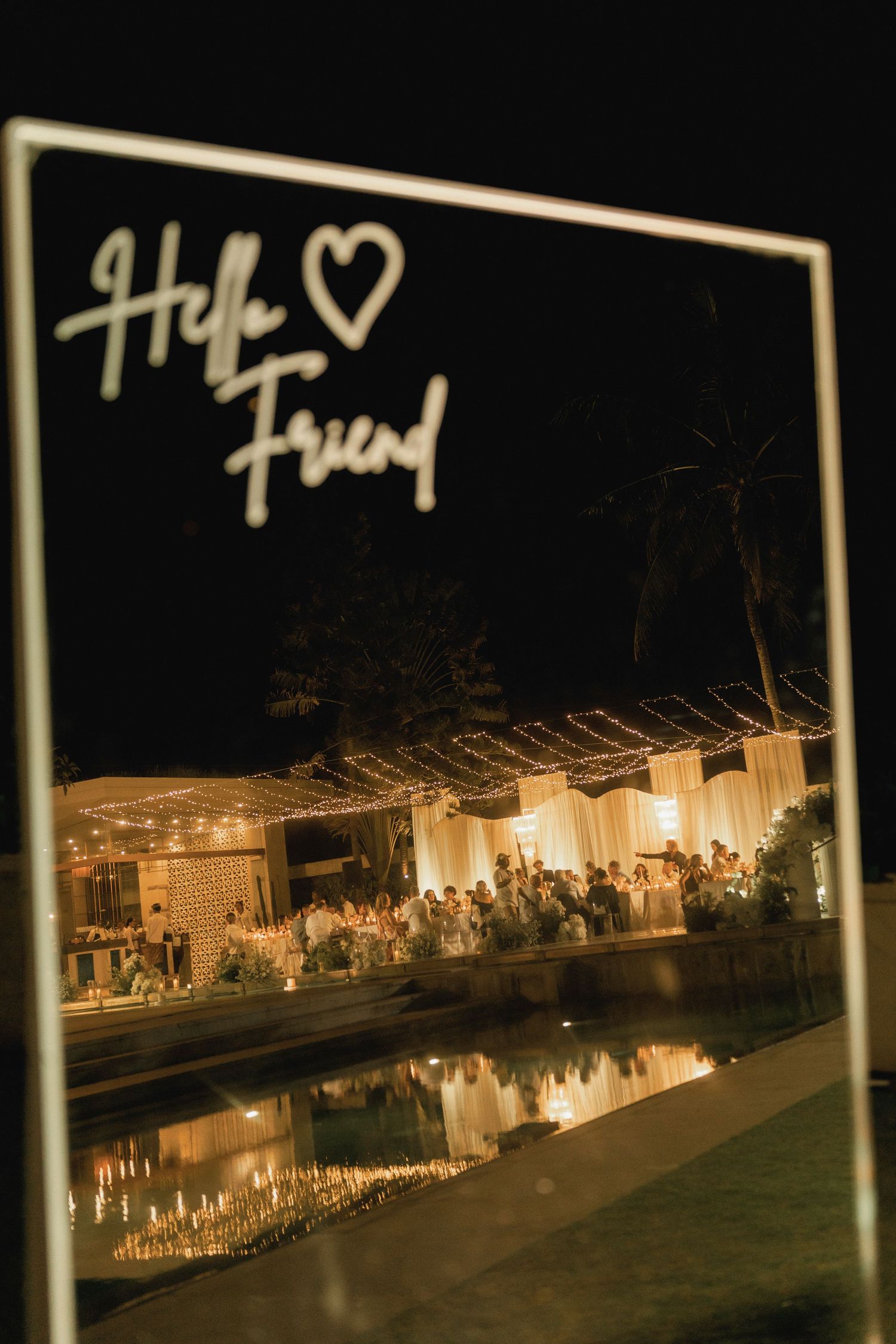Illuminated acrylic wedding welcome sign reading Hello Friend reflecting glowing outdoor reception venue at night.