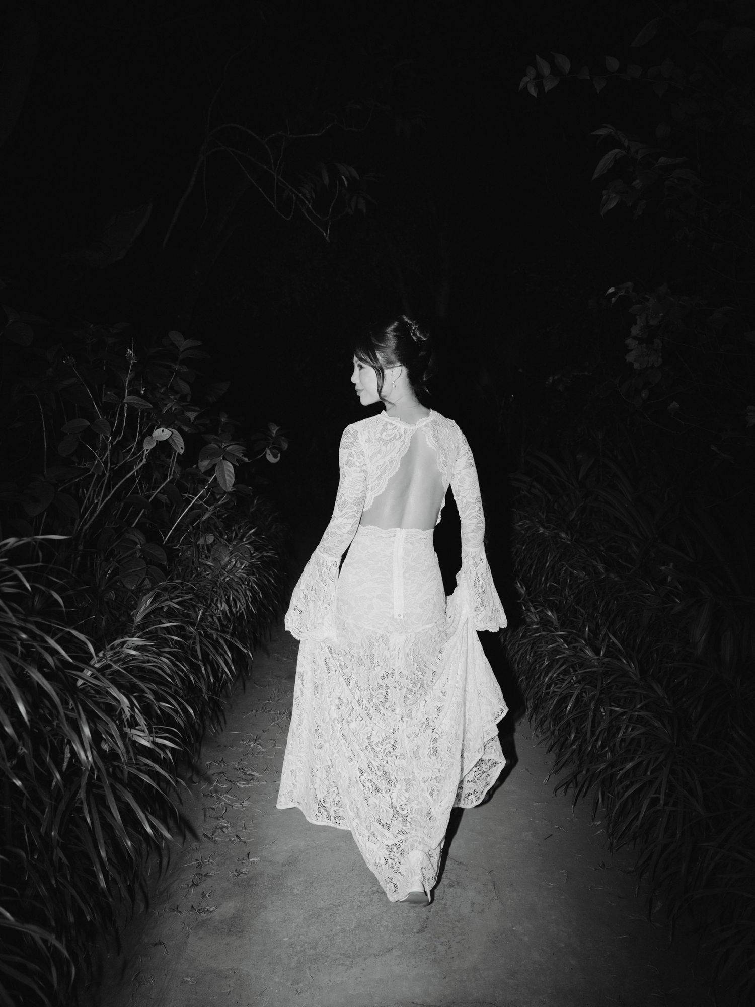 Bride in a backless lace wedding gown walks down a dark garden path at night.