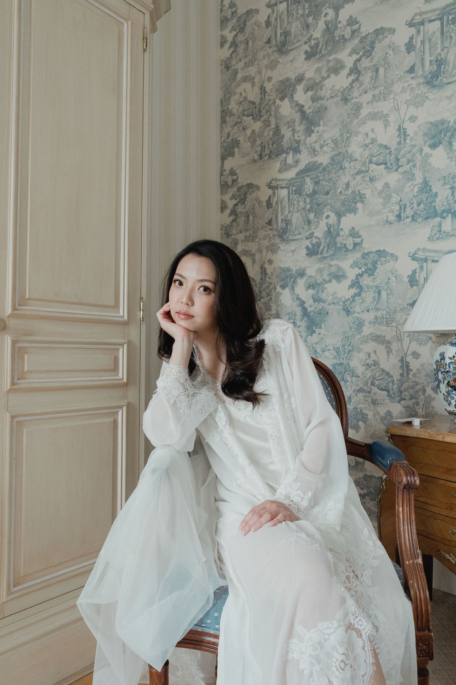 Bride in white lace robe sitting thoughtfully in a French-style room with toile de jouy wallpaper.