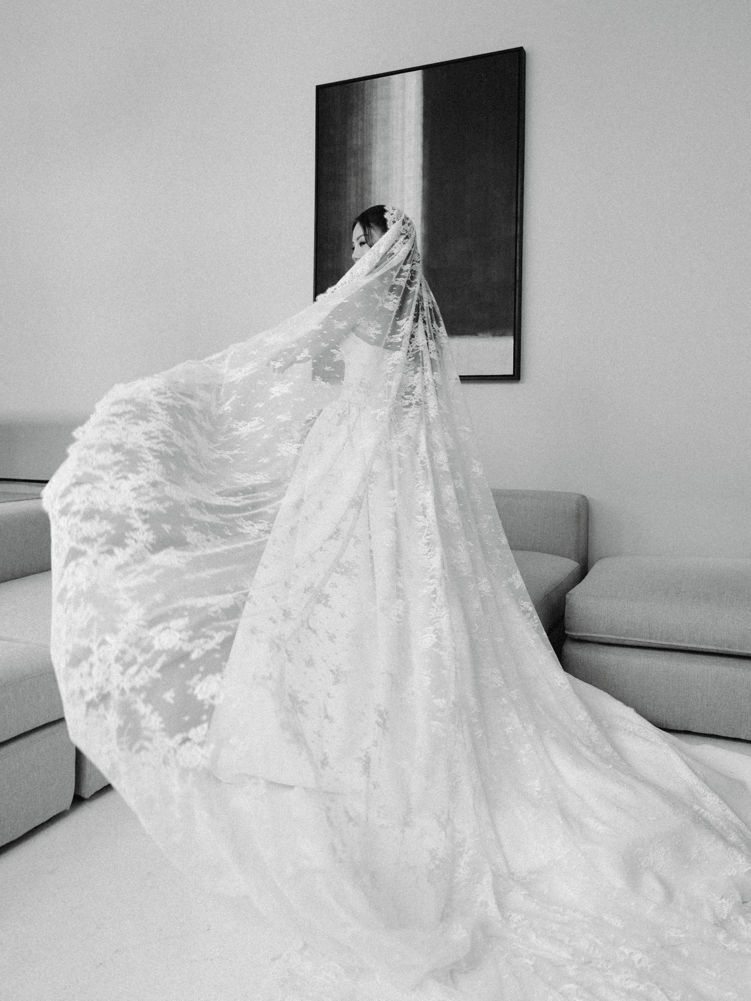 Black and white photo of bride in ornate lace cathedral veil spread dramatically across a modern room.