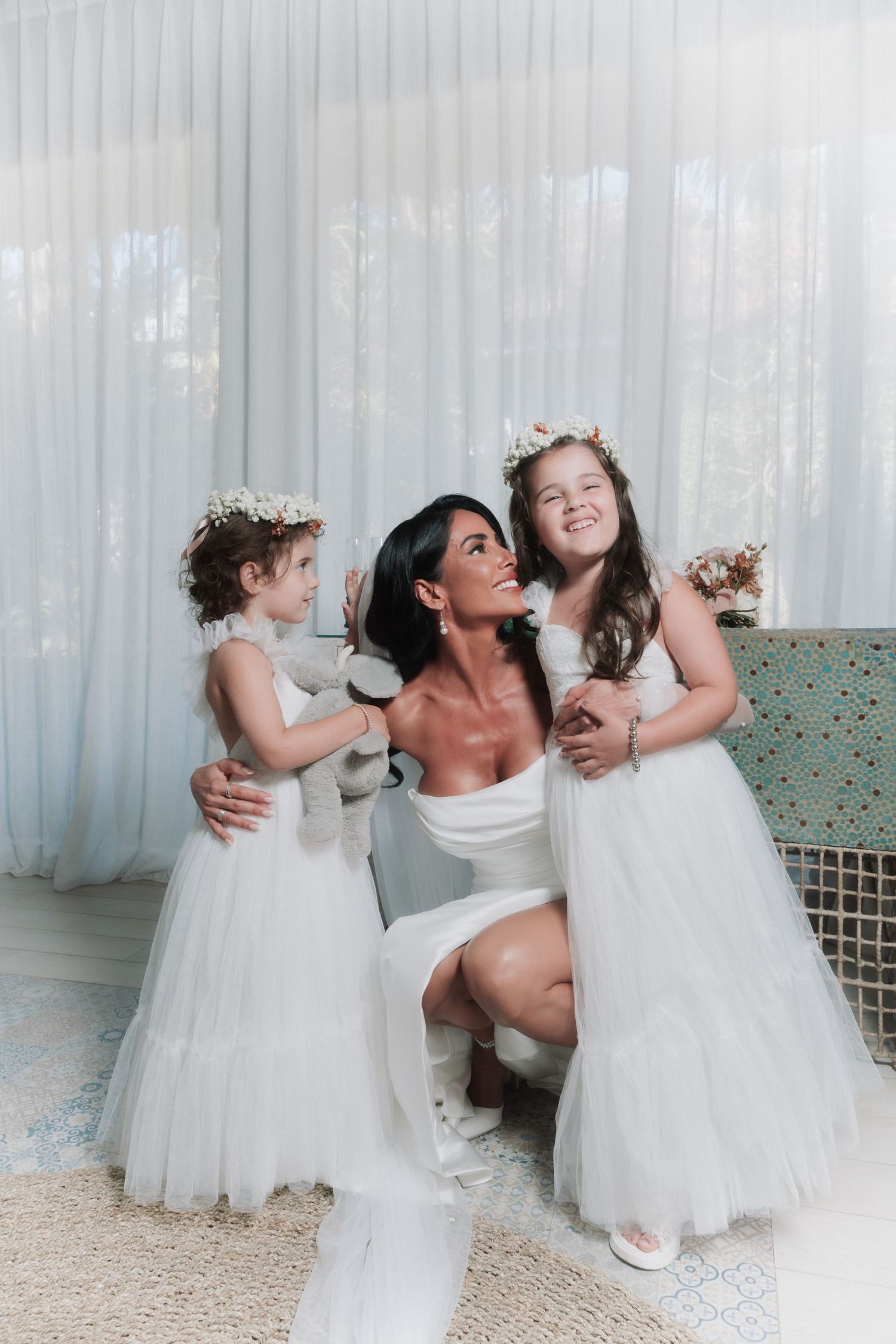 Bride in white dress kneeling with two flower girls in tulle gowns and floral crowns in a bright room.
