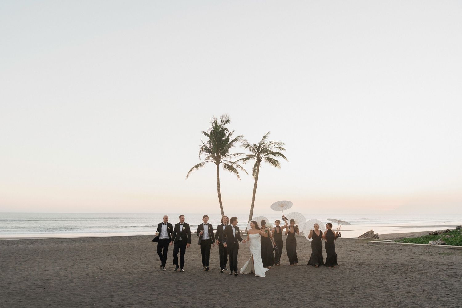 Wedding party walking along tropical beach at sunset with palm trees and ocean backdrop.