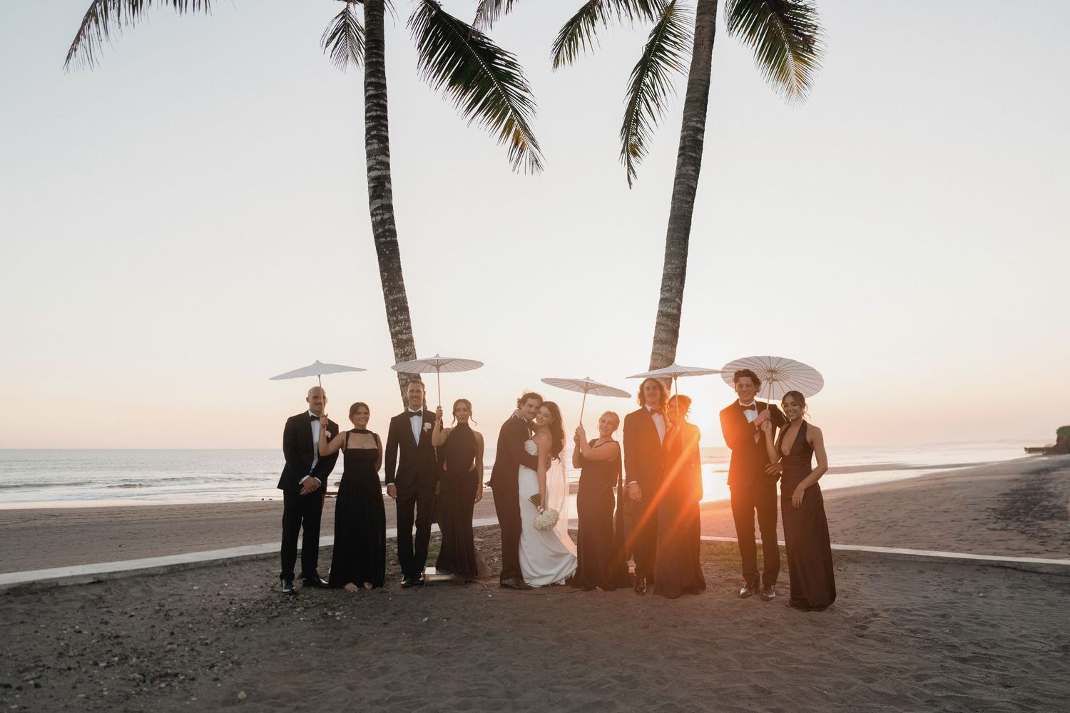 Wedding party posing at sunset beach with palm trees, bride and groom surrounded by guests in black attire.