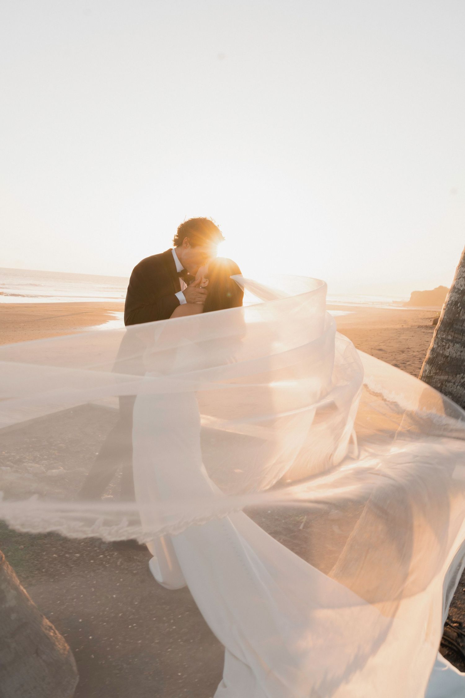 Bride and groom kissing at sunset on beach with flowing white veil billowing in the wind.