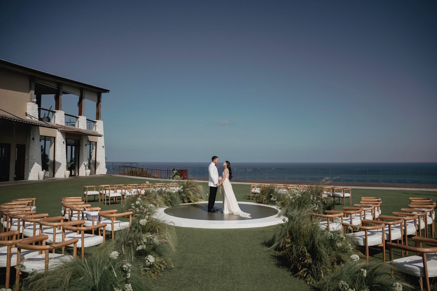 Bride and groom standing at outdoor ocean view wedding ceremony with wooden chairs and greenery.