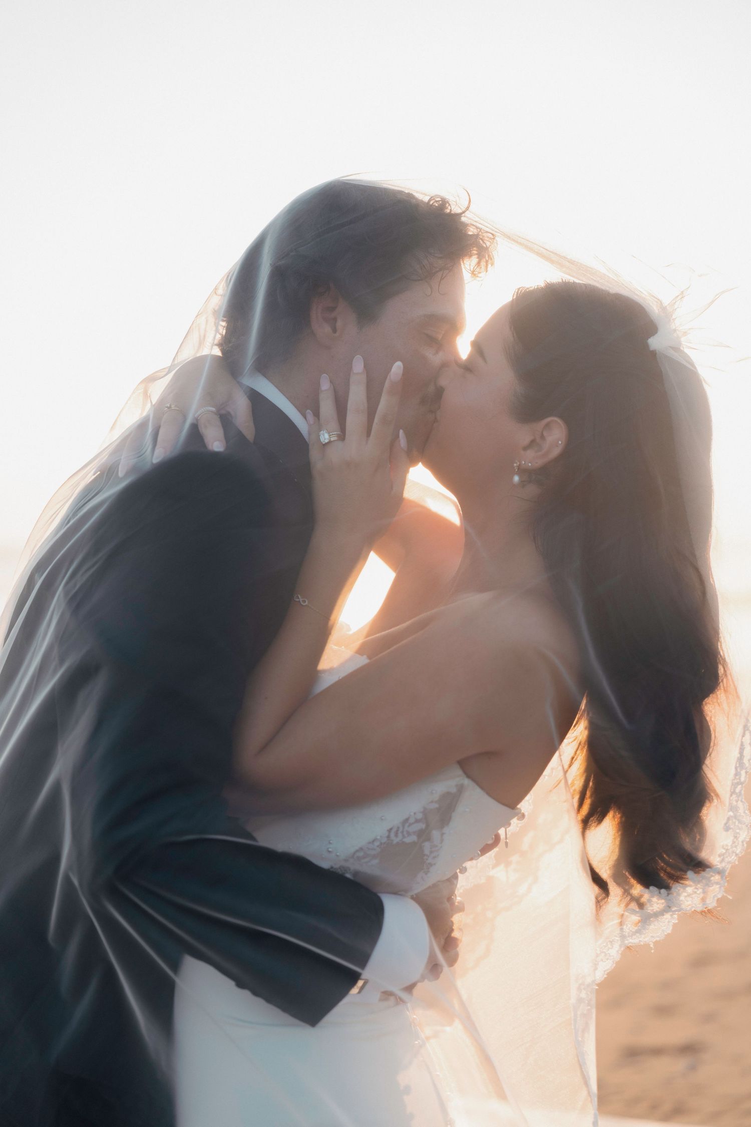 Bride and groom sharing romantic kiss under veil at sunset on beach wedding day.