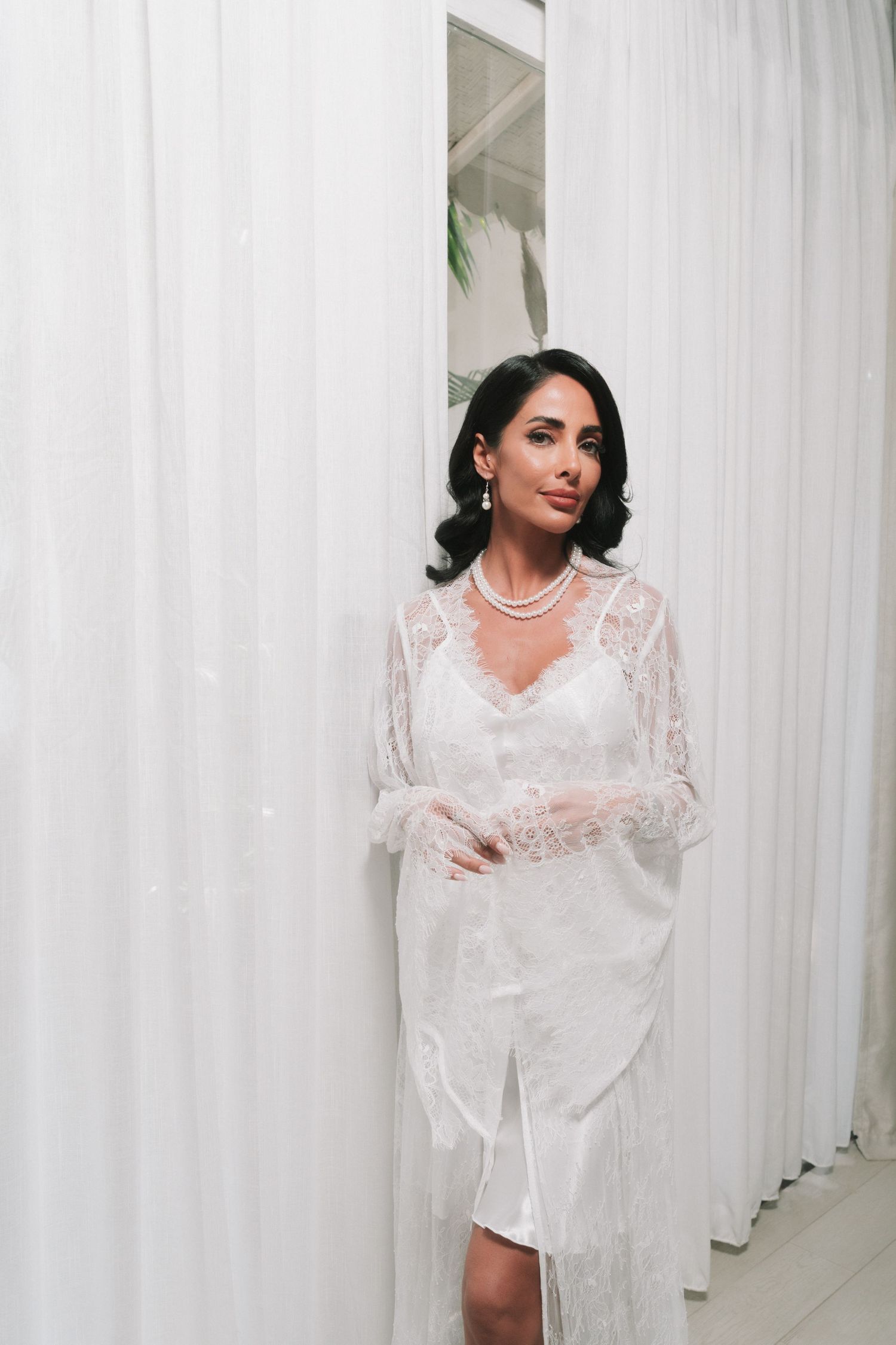 Elegant bride in lace robe and pearl necklace poses between sheer white curtains.