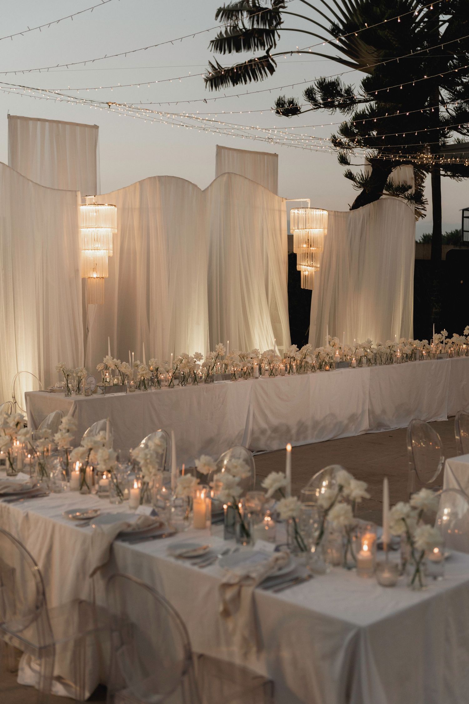Elegant outdoor wedding reception with white draped backdrop, chandelier lighting, and white floral centerpieces at dusk.