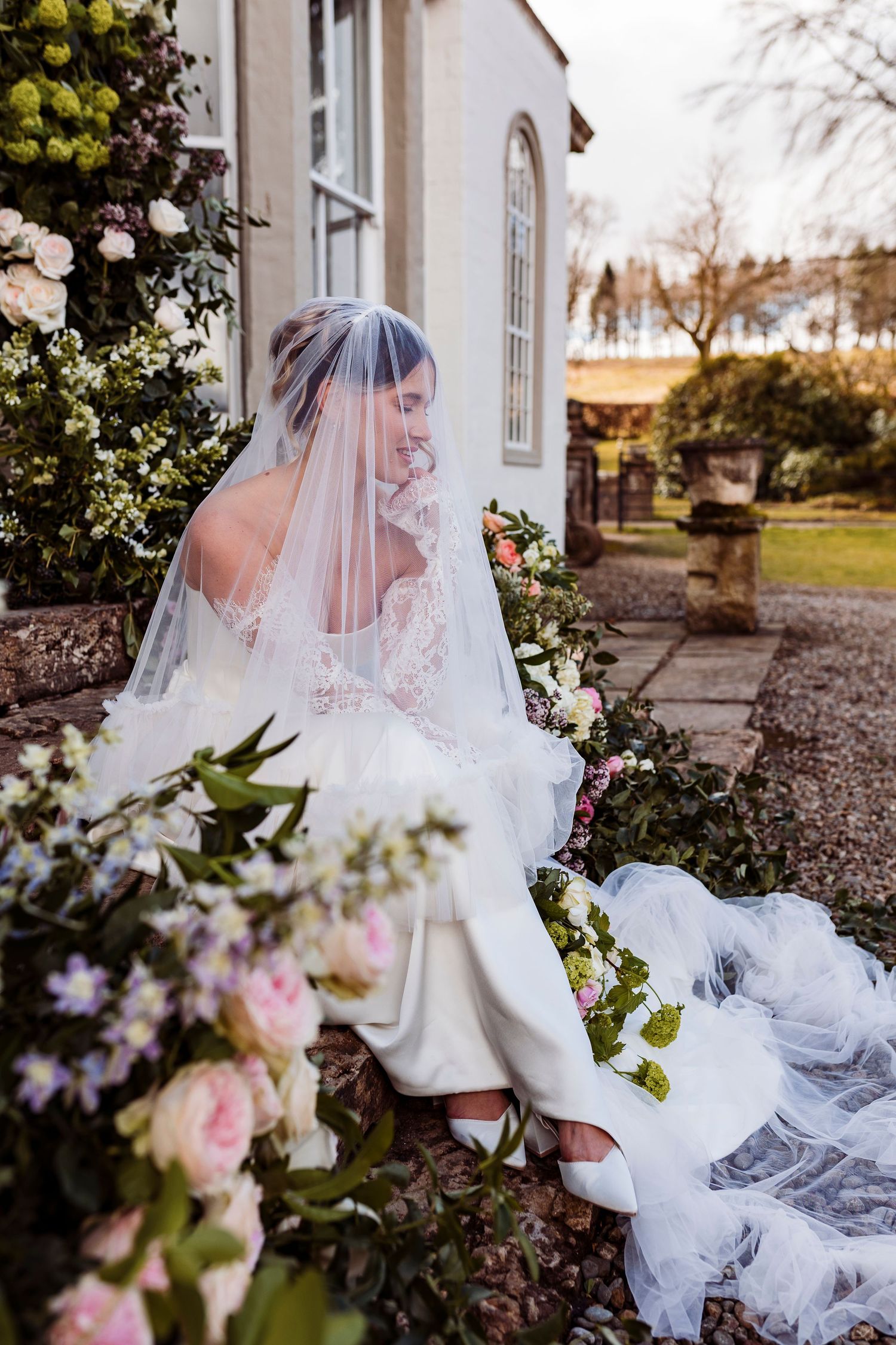 A bride in a flowing white dress and veil sits on stone steps surrounded by pink and white floral arrangements at a chapel.
