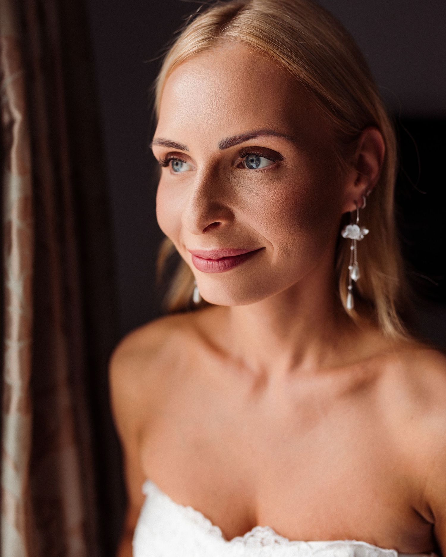 Bride in white strapless wedding dress smiles warmly in elegant indoor portrait with soft lighting.