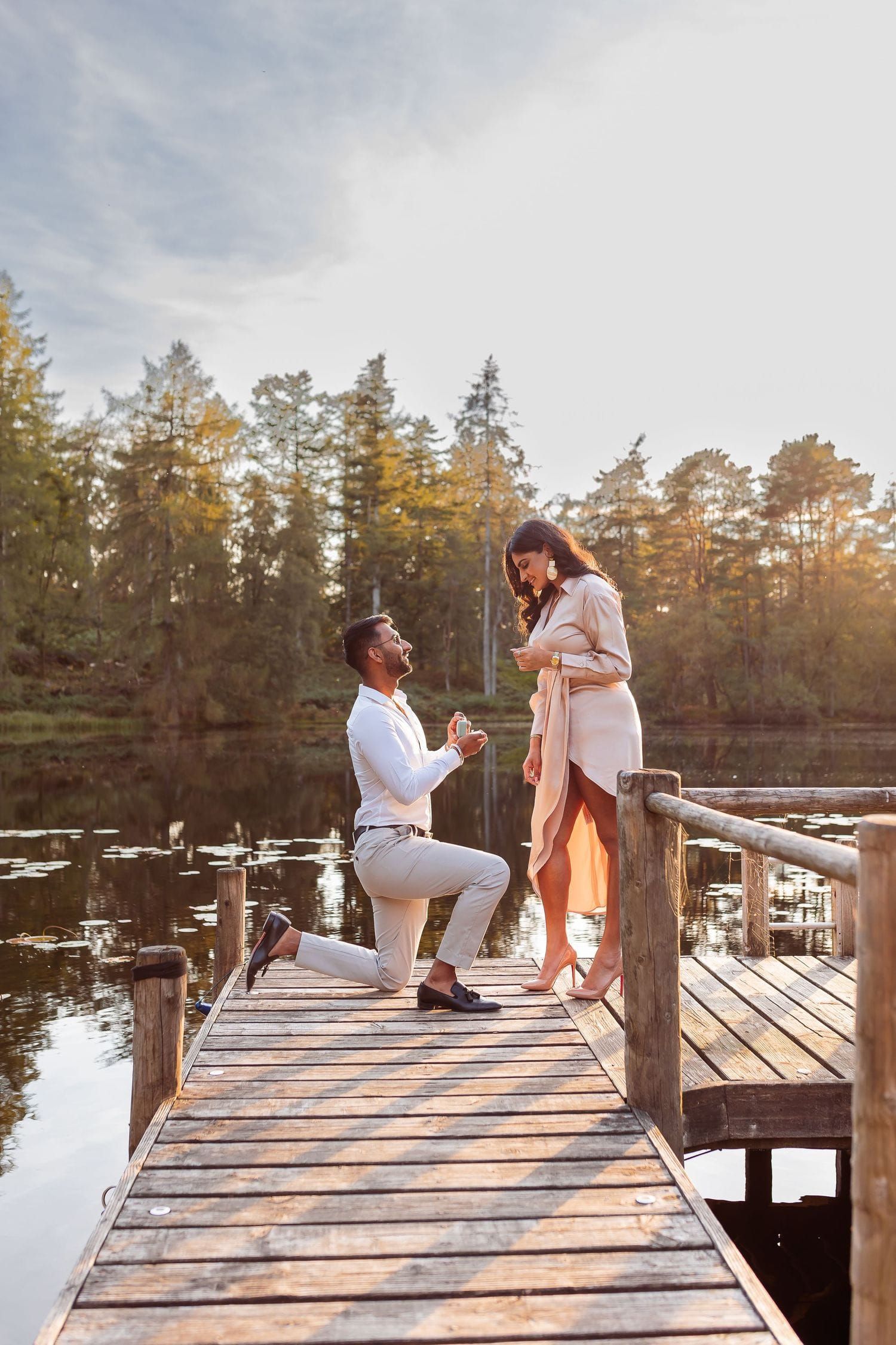 Man proposes to woman on wooden dock by lake surrounded by autumn forest at golden hour.