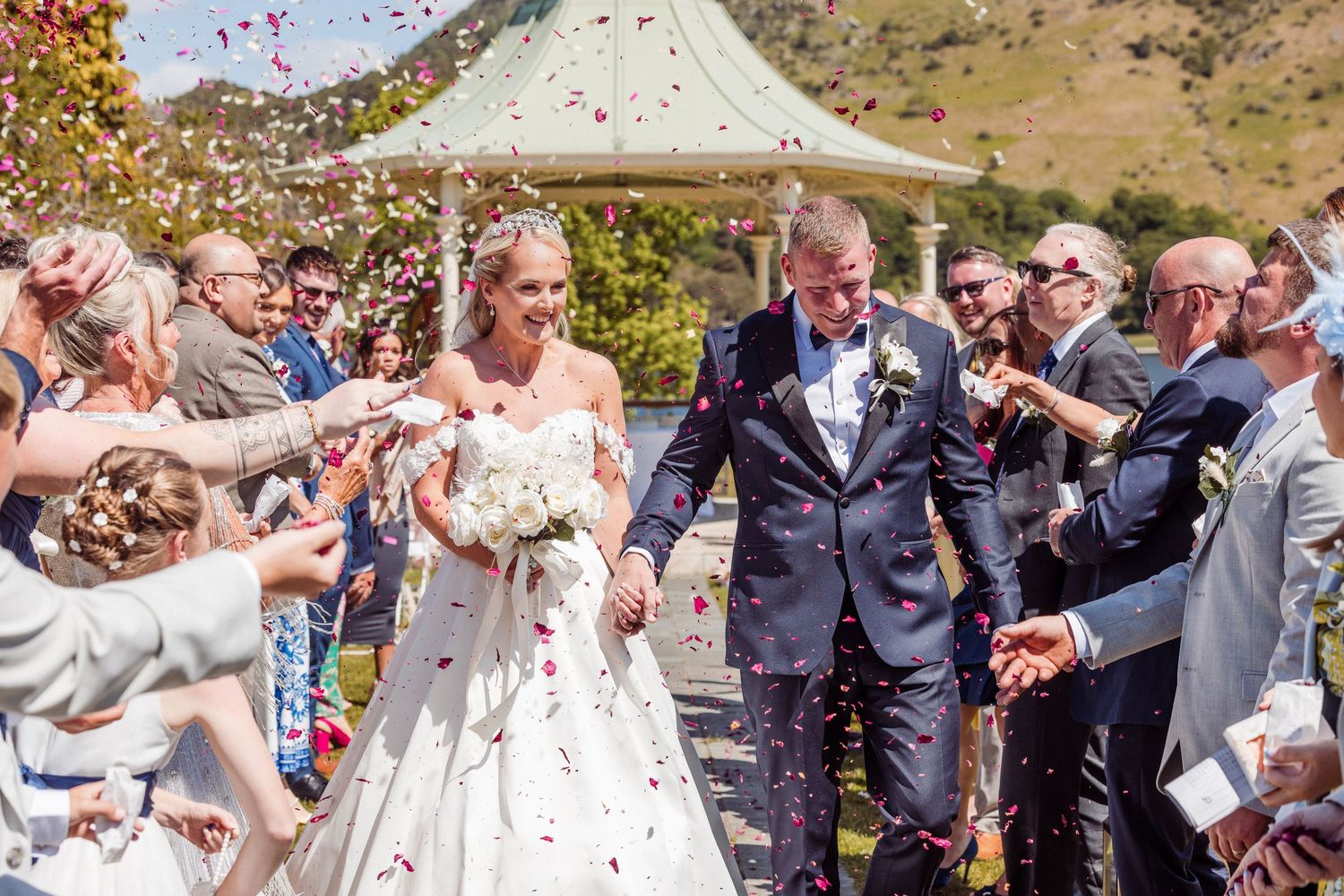 Bride and groom walk through confetti shower after outdoor wedding ceremony near a gazebo on a sunny day.