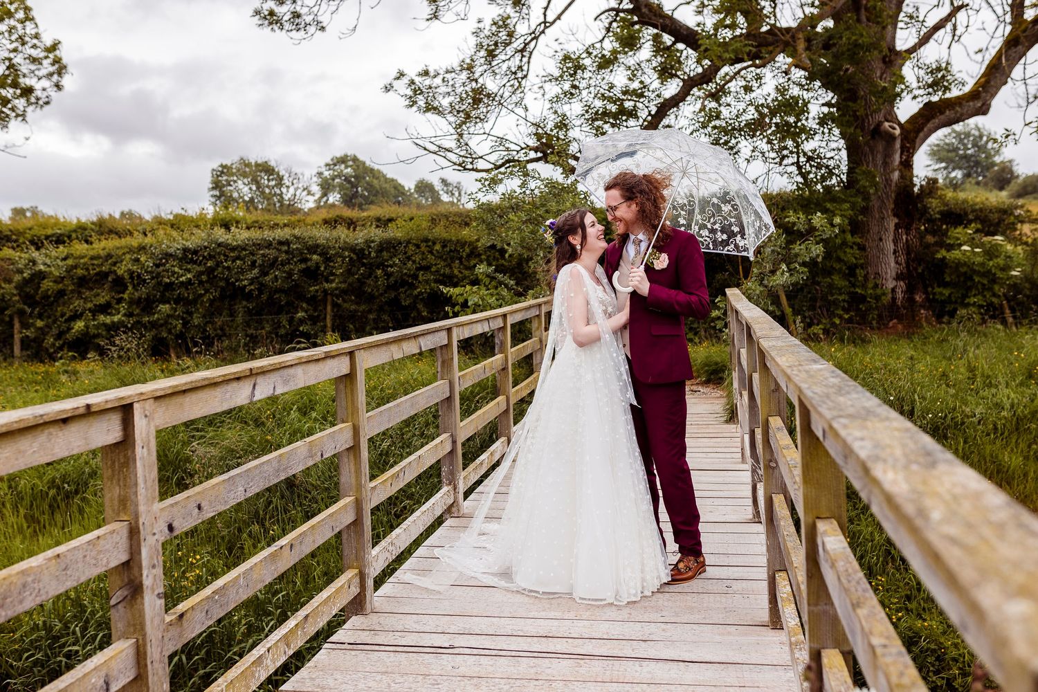 Bride and groom share a romantic moment under a clear umbrella on a wooden bridge surrounded by lush greenery.