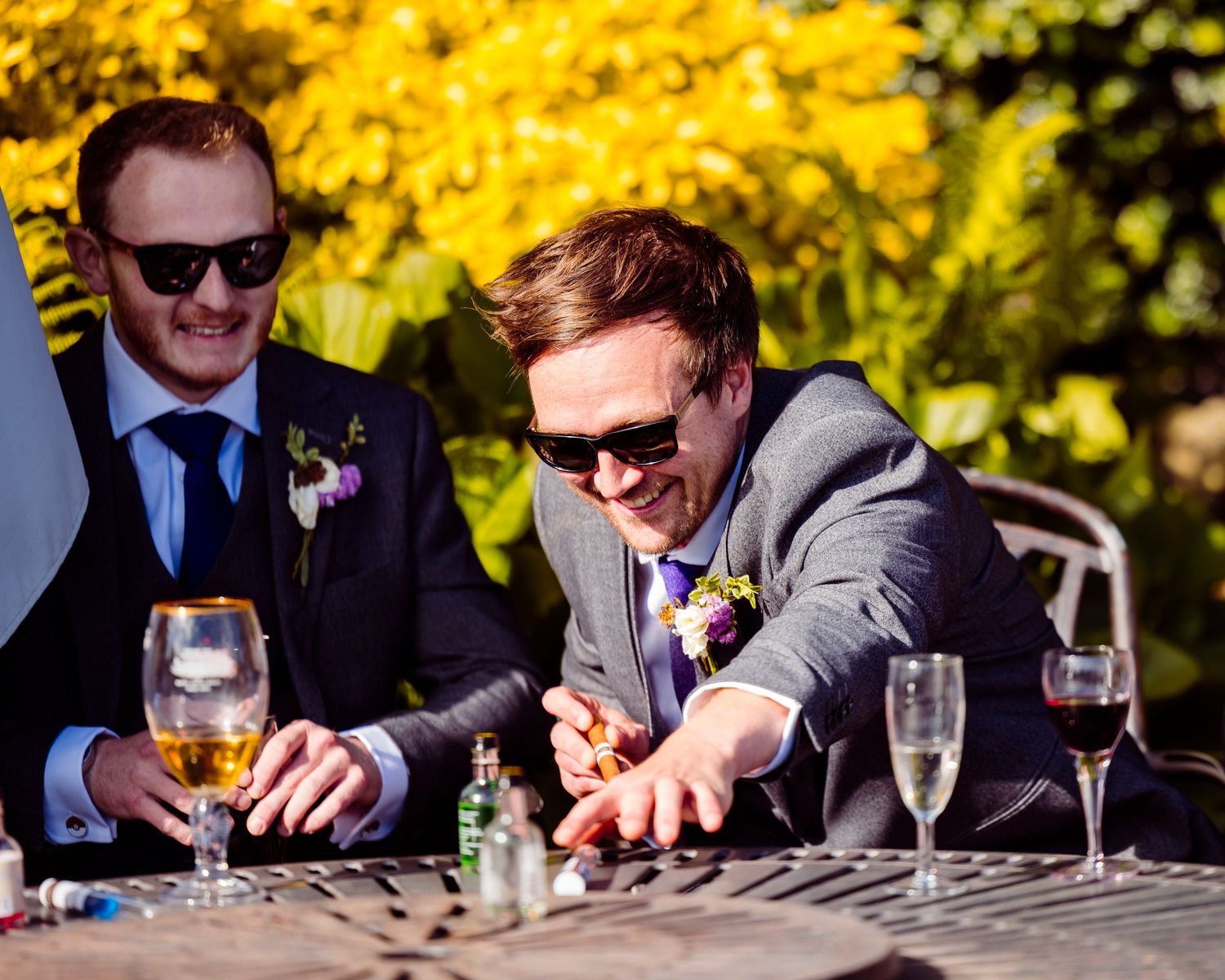 Two groomsmen in suits with boutonnieres laugh and play a game at an outdoor wedding reception table.