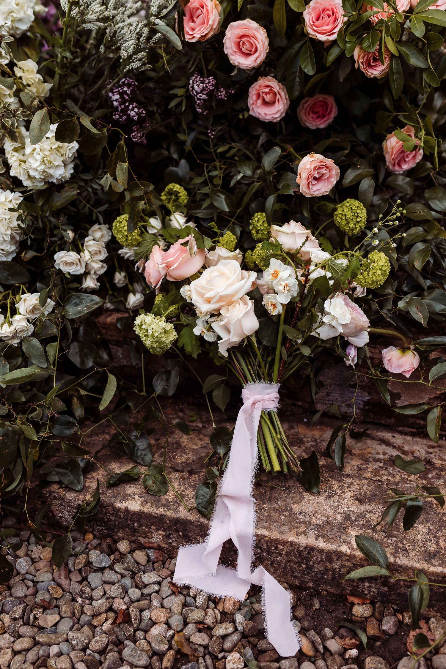 A romantic bridal bouquet with pink roses and white flowers tied with a silk ribbon rests on stone steps among lush greenery.