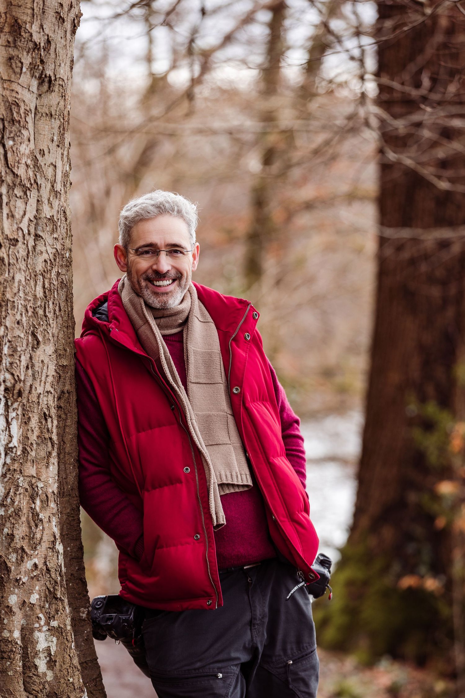 Middle-aged man with gray hair smiling in red vest and scarf, leaning against tree in autumn forest.