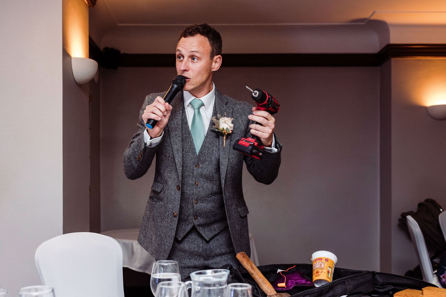 Man in grey suit giving a speech at a wedding reception, holding a microphone and a drill as a prop.