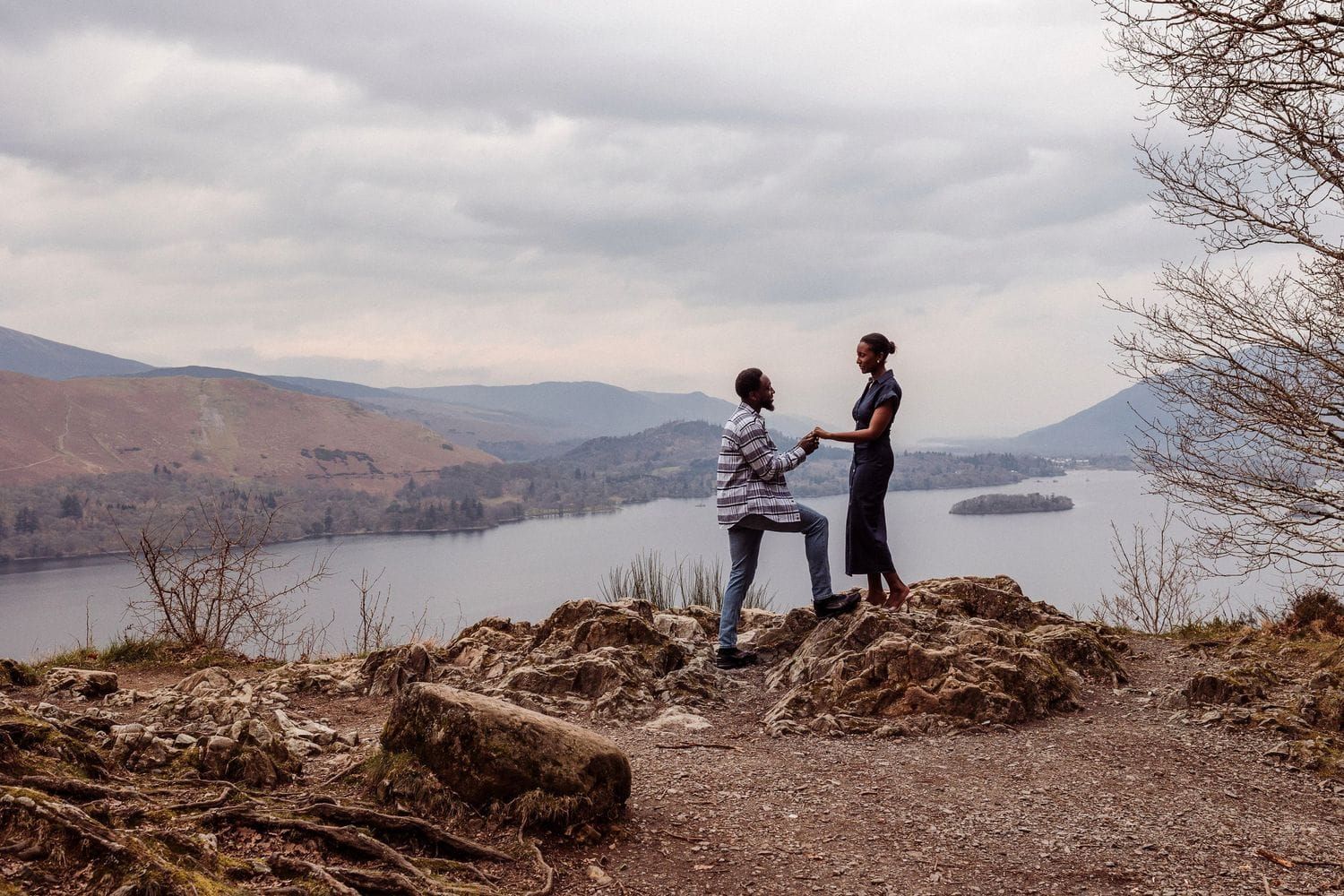 Two people stand on a rocky hilltop at Surprise View overlooking Derwentwater and mountain views under cloudy skies.