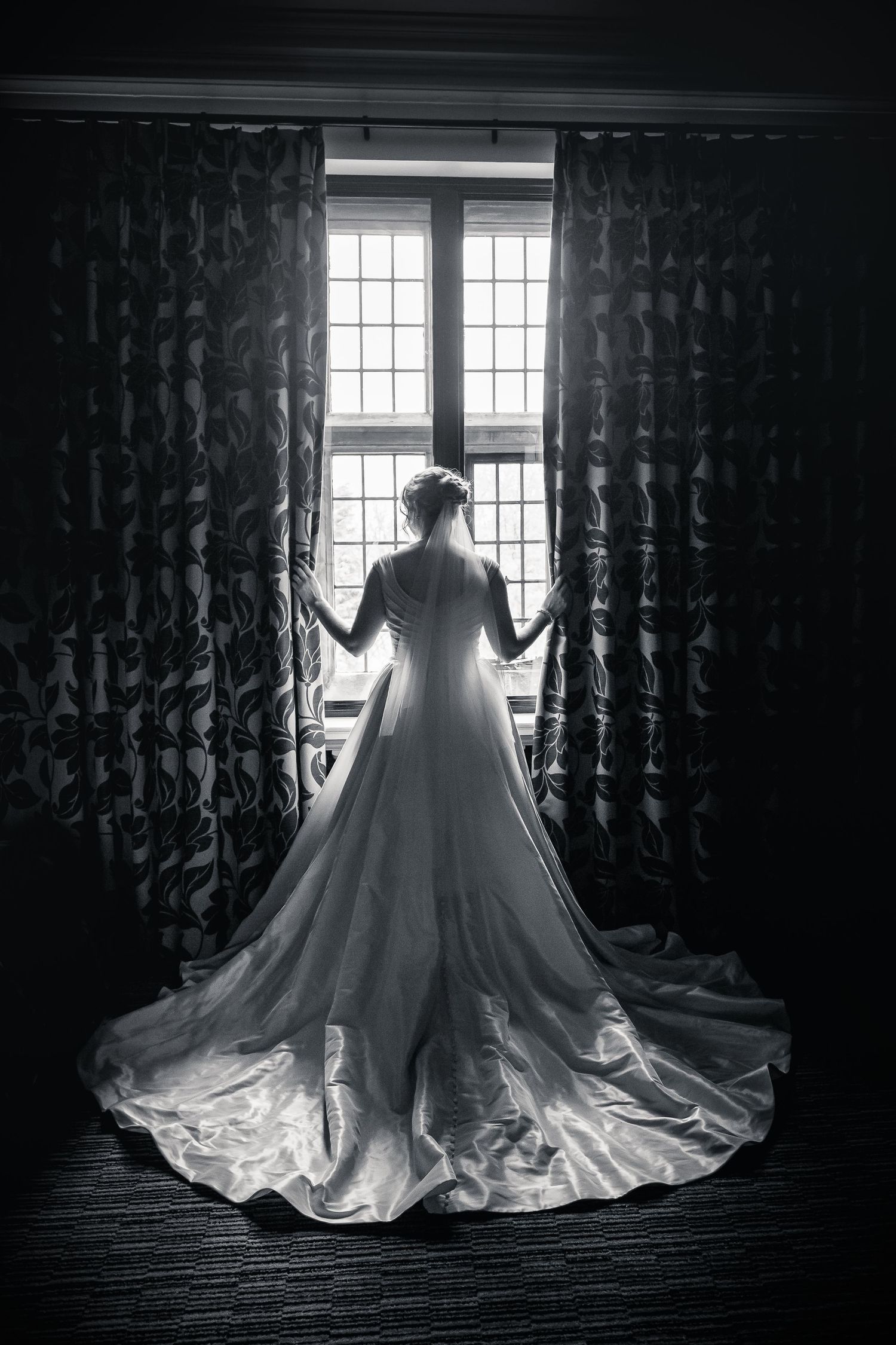 Dramatic silhouette of a wedding dress with flowing train against backlit windows and dark curtains in black and white.