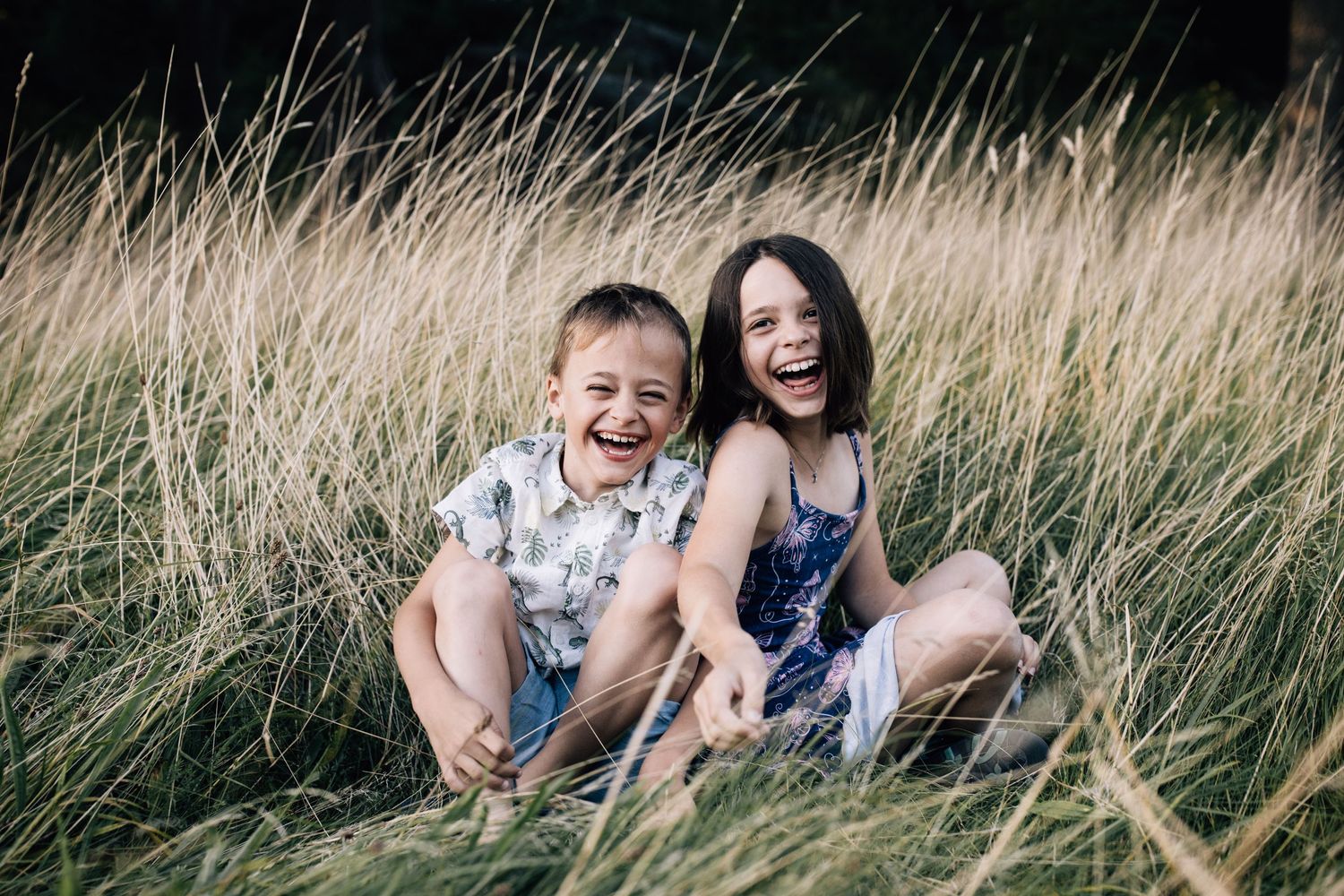 Two children laughing together while sitting in tall grass on a sunny day.