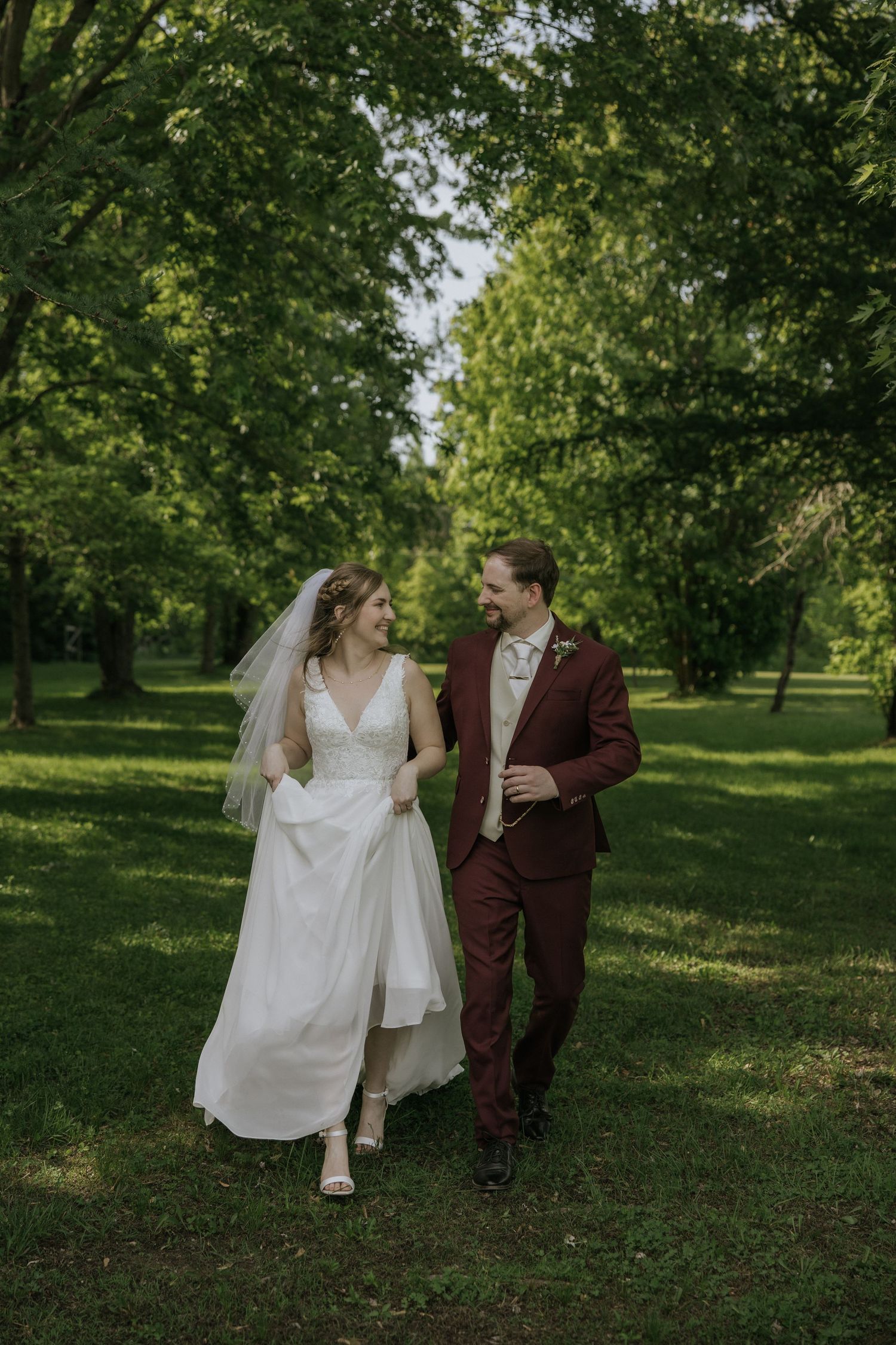 A couple in wedding attire walks through a lush green park setting in spring, with flowing white dress and burgundy suit.